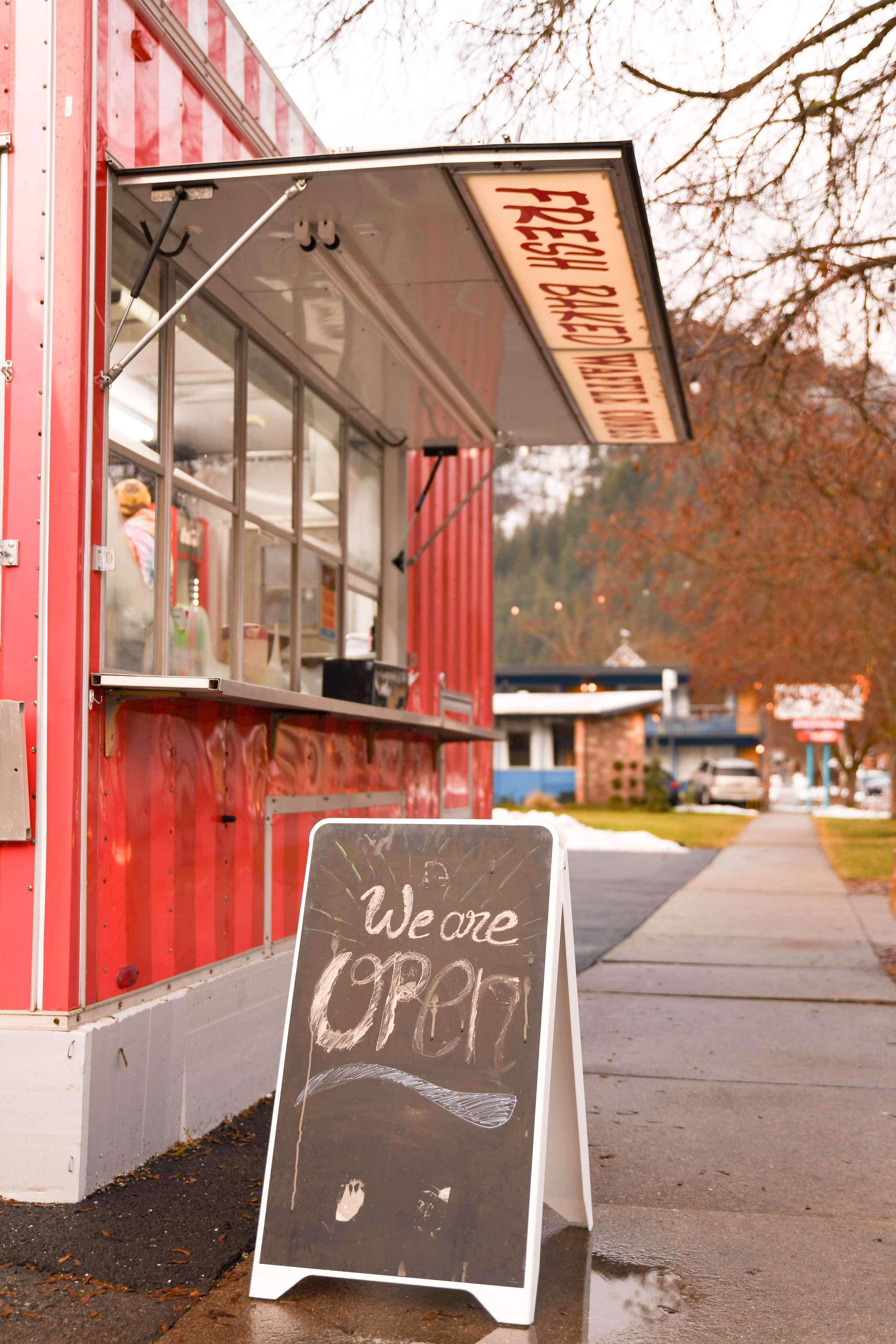 Red food stand with a sign that says "We are Craf" and a smaller sign above it that says "Open 11AM - 9PM"; sidewalk with trees and houses in the background.