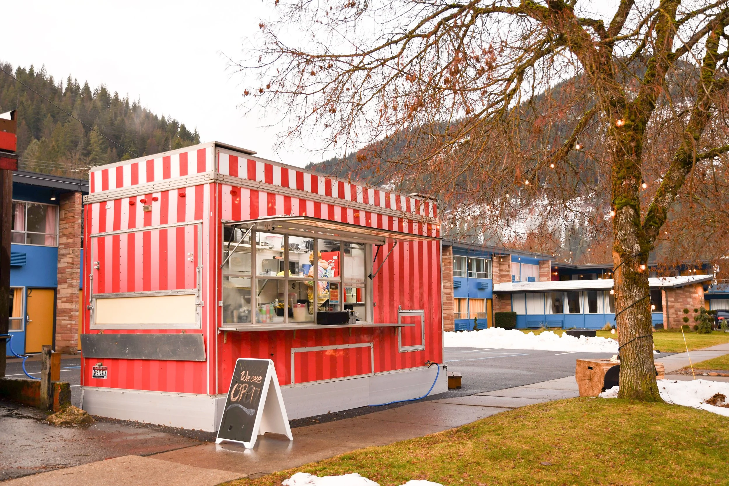 Red striped food truck with a window, located in a residential area with snow patches on the ground. There is a chalkboard sign in front of the truck that says "We are OPEN." A large tree with string lights is nearby, and apartments with colorful fac