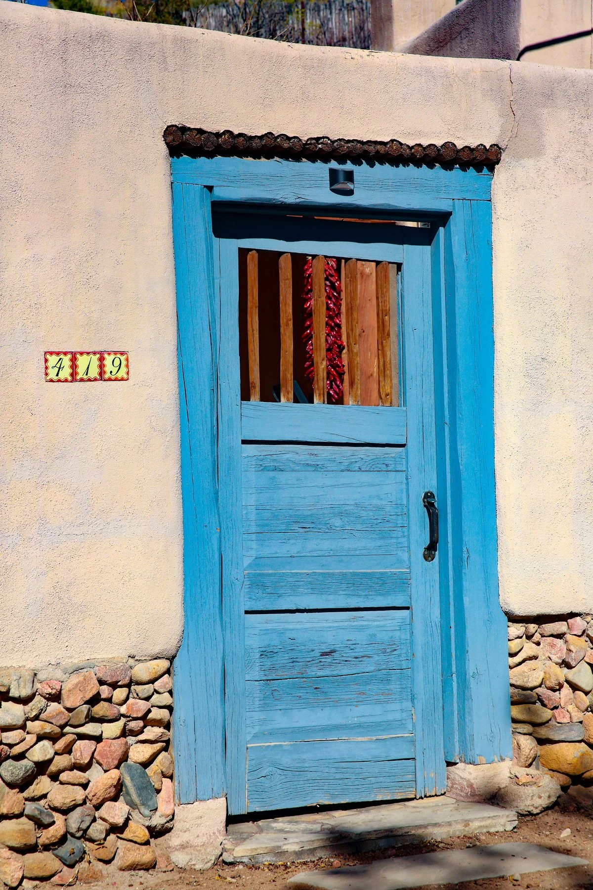 Blue wooden door with black handle set in a beige stucco wall with pile of rocks at the bottom, house number 419 on a small sign to the left, and a red chili pepper hanging inside visible through the top window.