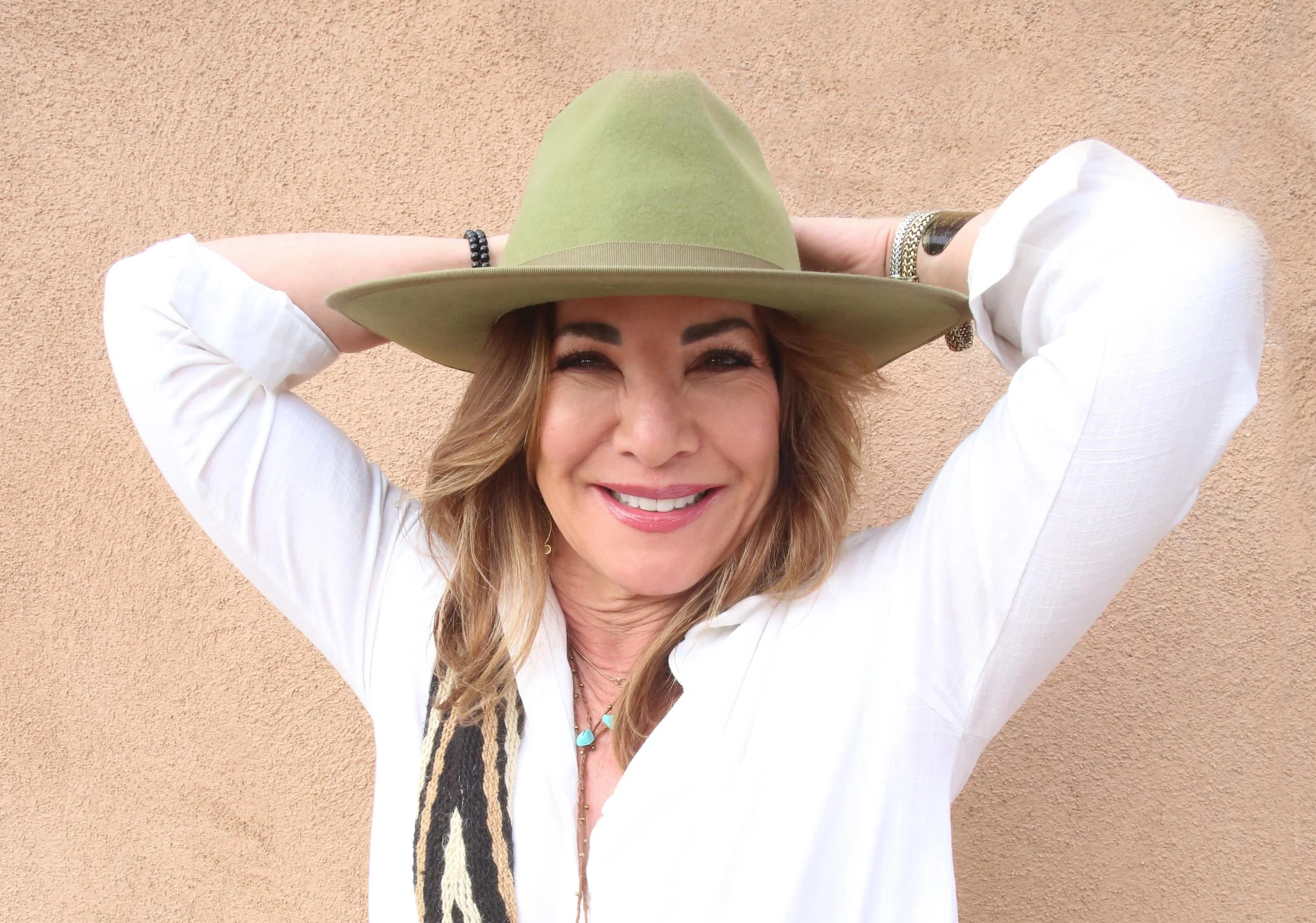 A woman smiling, wearing a large green hat and white shirt, standing in front of a peach-colored wall.