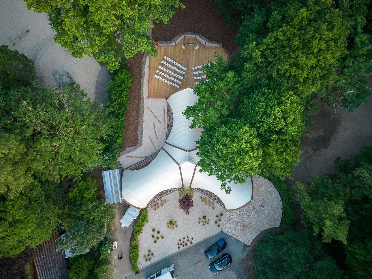 Aerial view of a pavilion with a unique, white, tent-like roof surrounded by lush green trees, a paved area with parking spaces, and a smaller gravel area with plants.