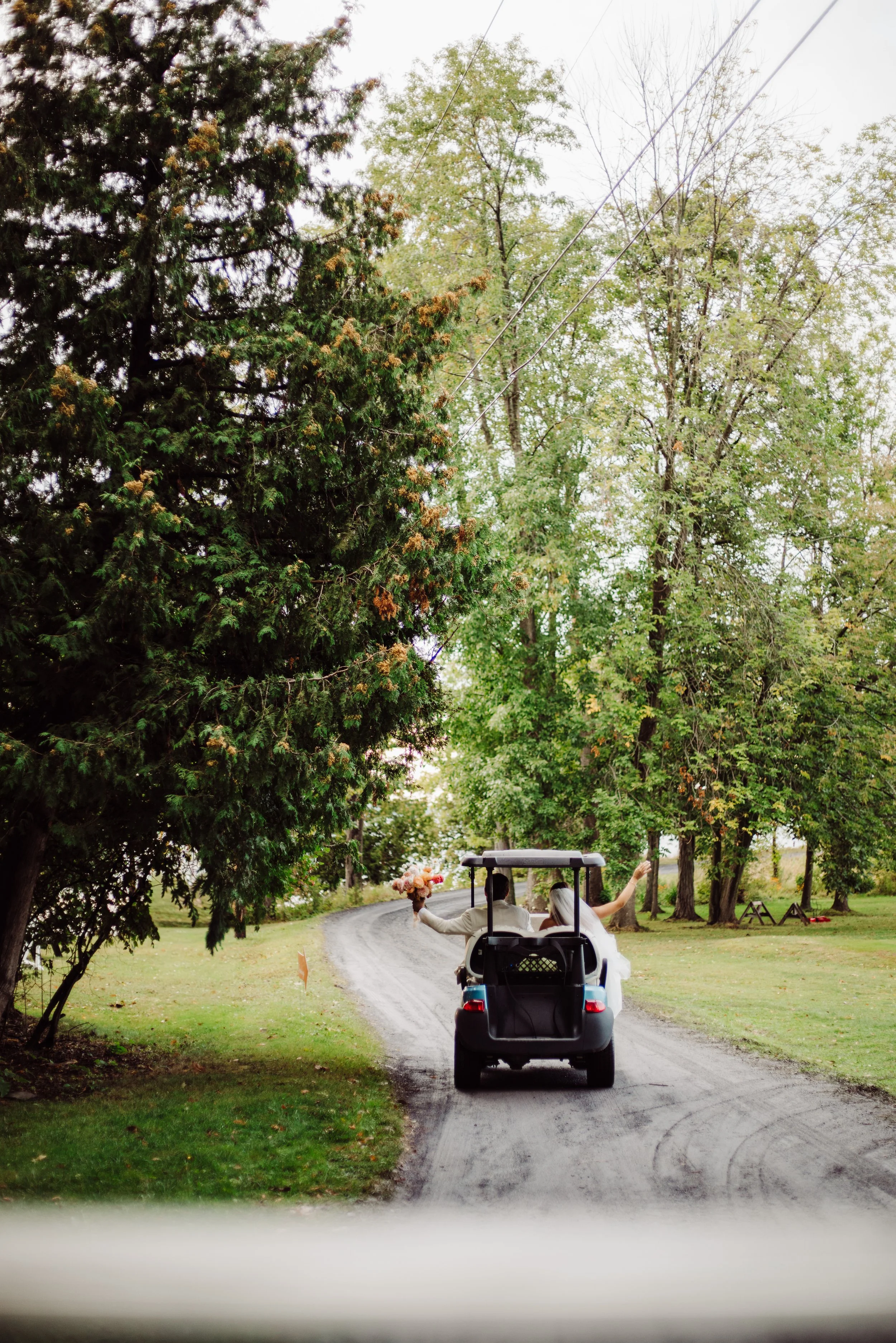 A golf cart with a bride and groom dressed in wedding attire, one holding a bouquet, driving down a dirt road lined with trees.