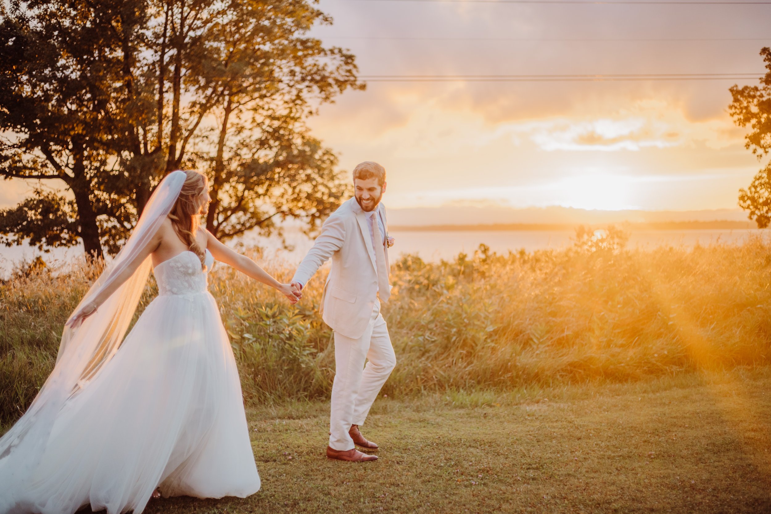 A bride and groom holding hands and walking outdoors during sunset, with trees and open sky in the background.