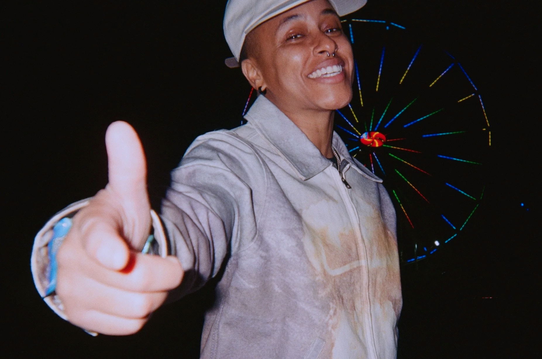 A person smiling and giving a thumbs-up at night with a colorful Ferris wheel in the background.