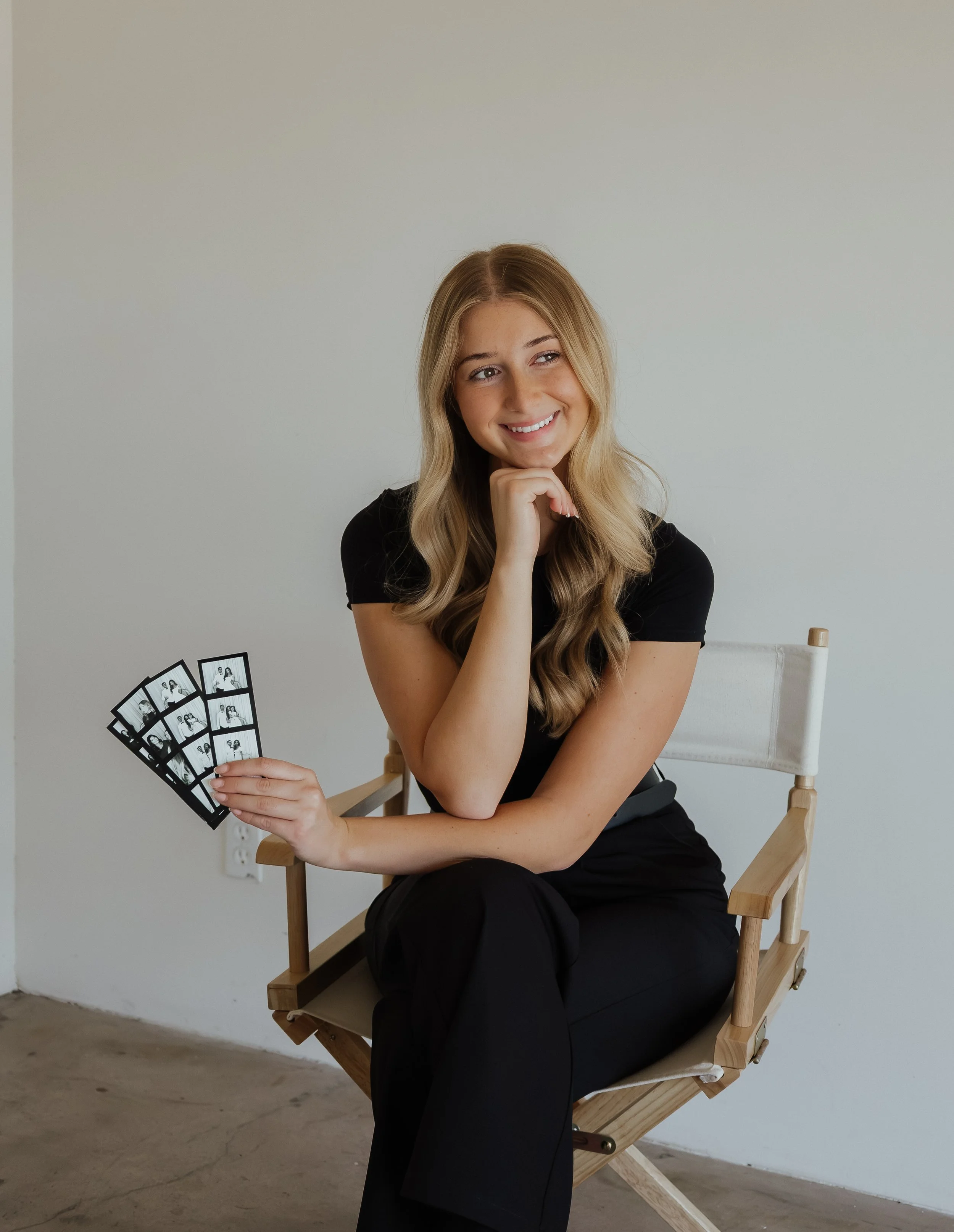 A young woman with long wavy blonde hair, wearing a black shirt and black pants, sitting on a wooden director's chair. She is smiling and holding four photo booth strips in her left hand, resting her chin on her right fist.