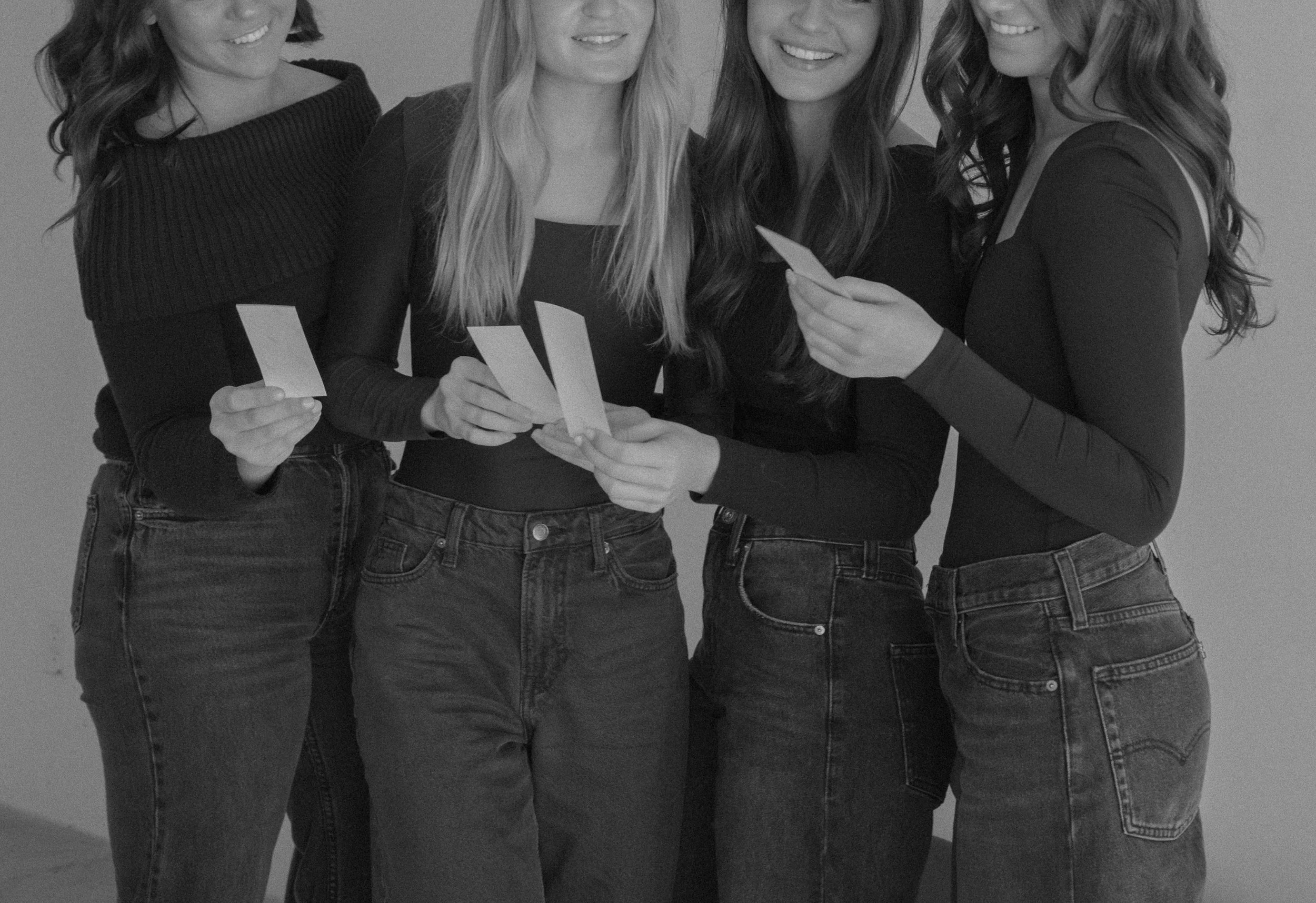 Four women standing together, looking at photographs or cards, in casual clothing and jeans