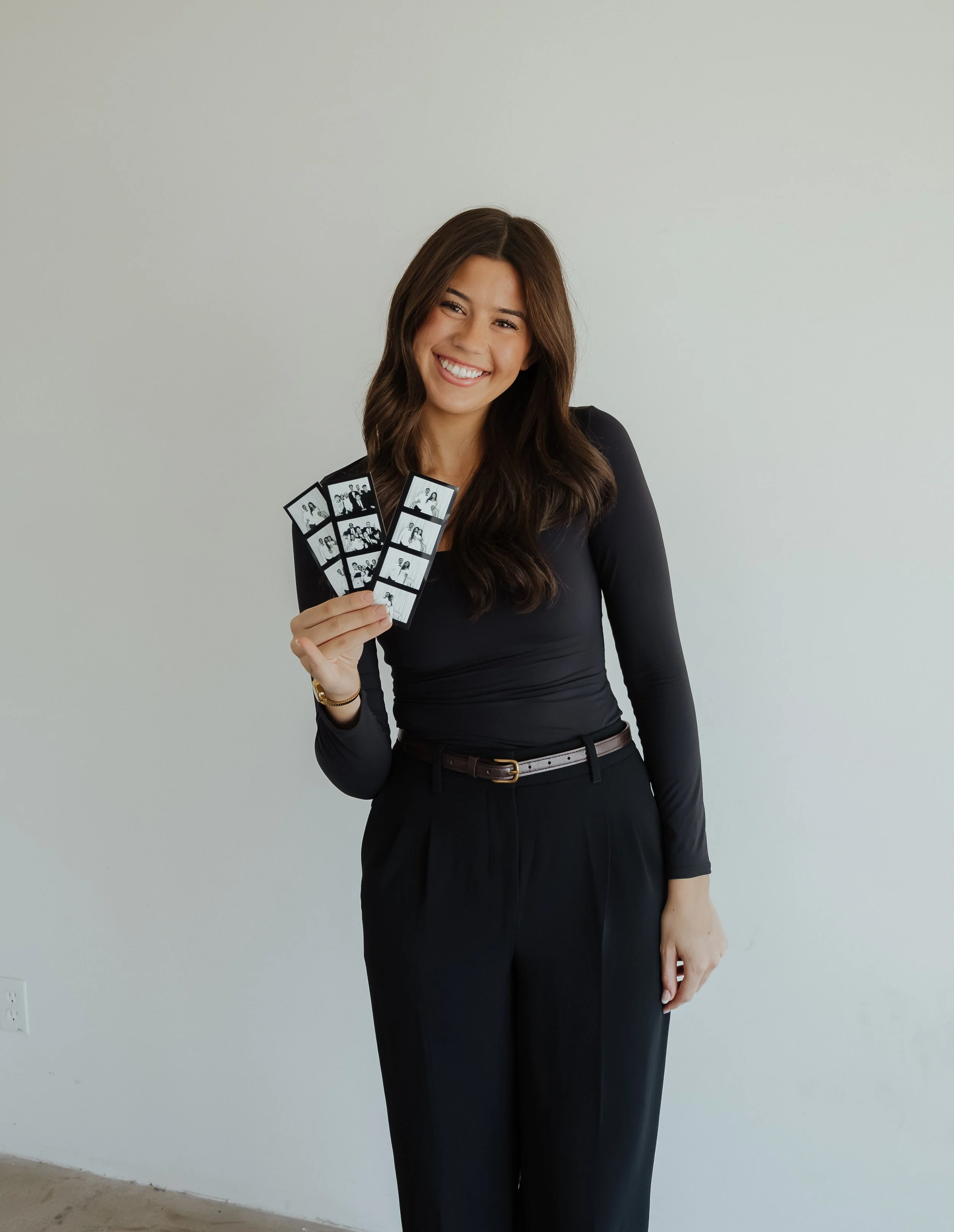 A woman in a black long-sleeve top and black pants holding photo booth strips, smiling in front of a plain wall.