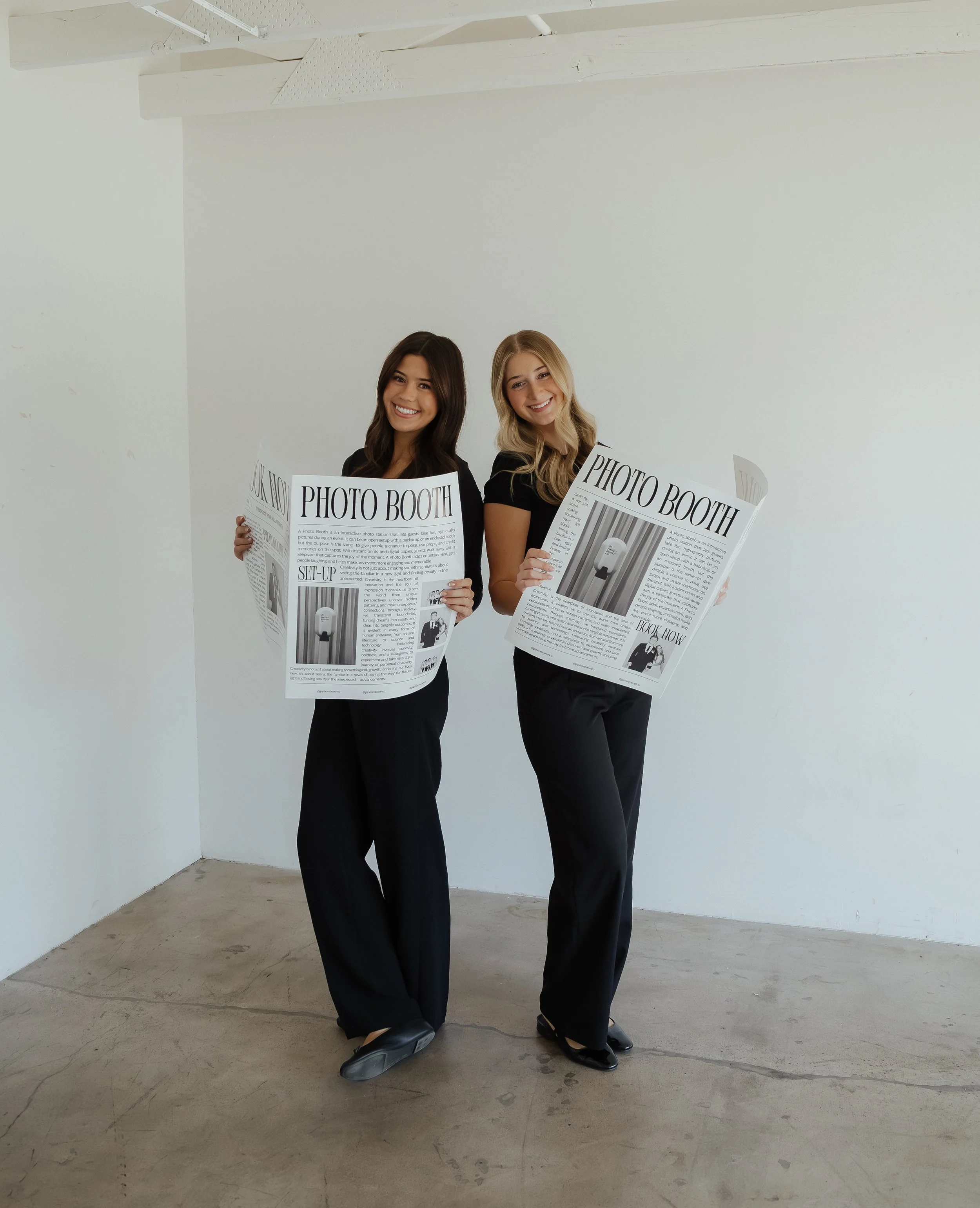 Two young female entrepreneurs holding newspapers with headlines about their photo booth, standing in a minimalistic room with white walls and concrete floor.