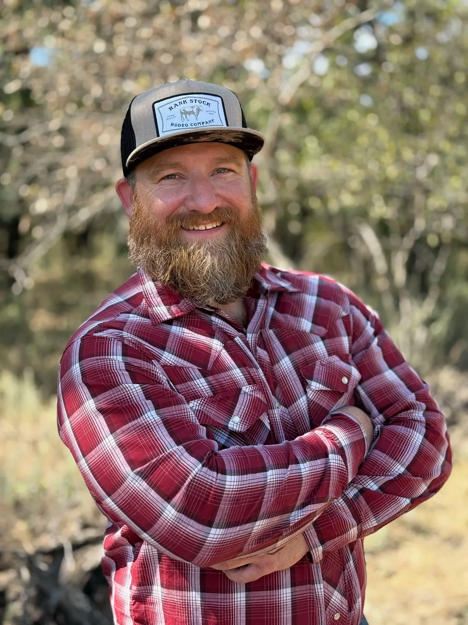 A man with a beard and mustache smiling, wearing a gray and black trucker hat with a logo, and a red plaid shirt with rolled-up sleeves, standing outdoors in a wooded area.