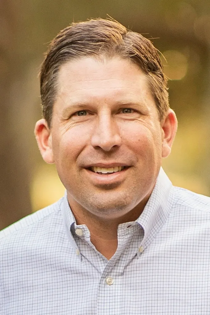 A smiling man with brown hair, wearing a light-colored, checkered button-down shirt, outdoors with blurred greenery in the background.