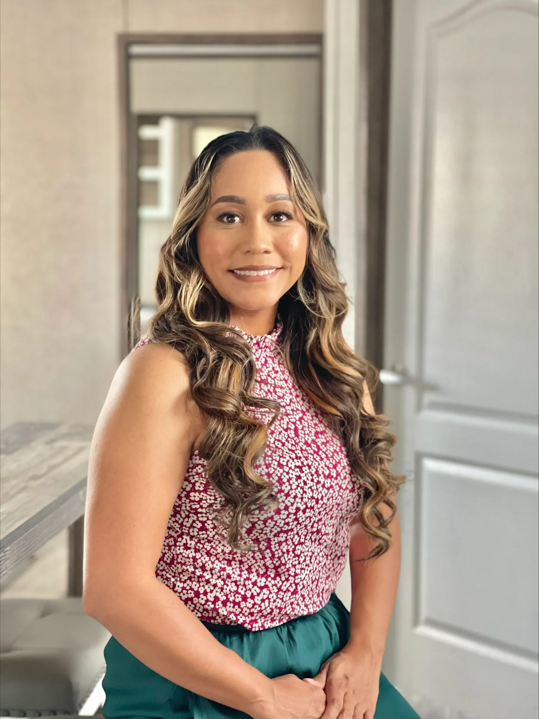 A woman with long, curly, brown hair styled with blonde highlights, wearing a sleeveless red and white floral top, sitting at a desk in a modern room with a mirror in the background.