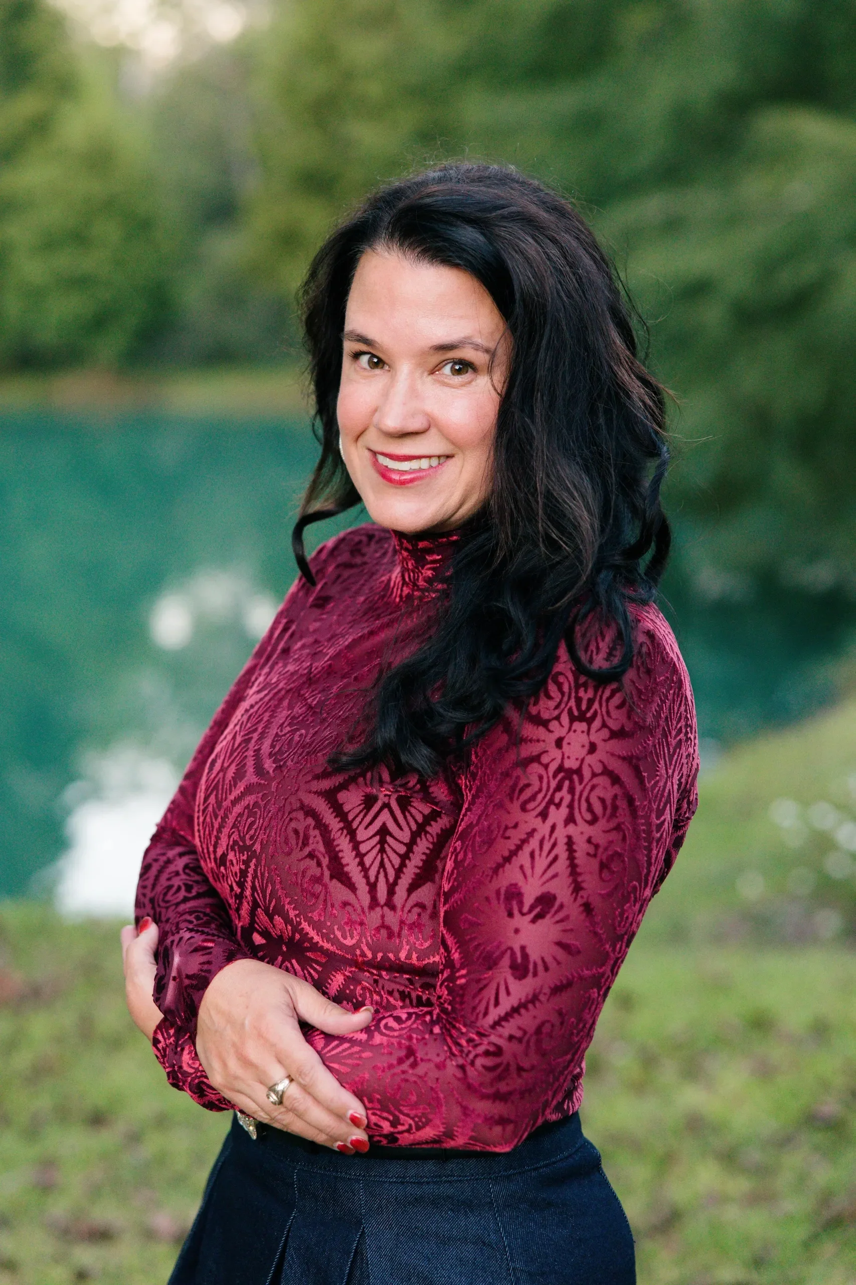 A woman with dark hair wearing a red patterned top stands outdoors near a lake with green trees in the background, smiling at the camera.
