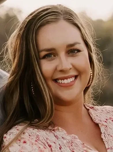 A smiling woman with long brown hair, wearing hoop earrings, outdoors in natural light.
