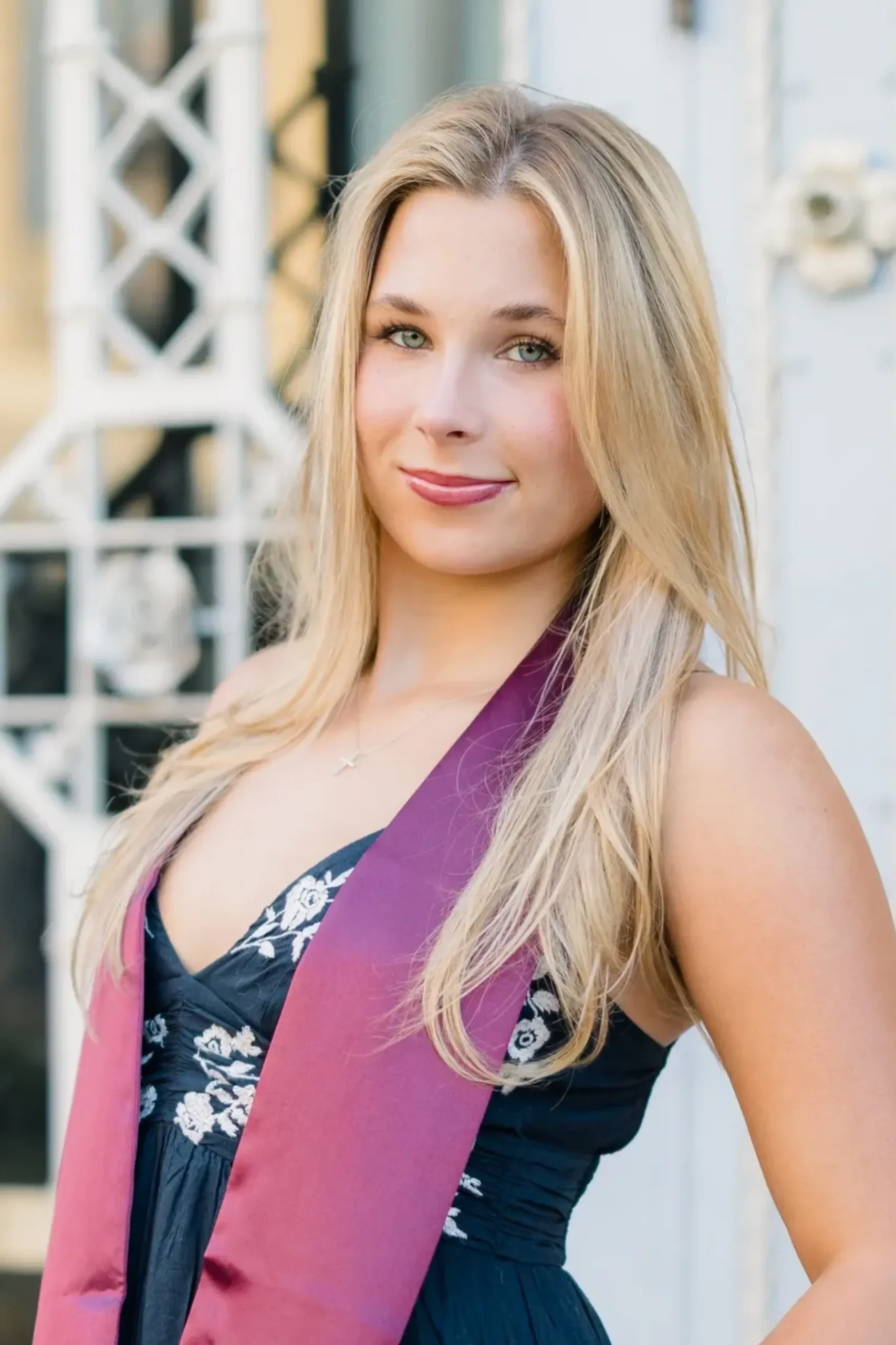 Young woman with long blonde hair wearing a graduation stole and dress, smiling outdoors with a blurred white structure in the background.