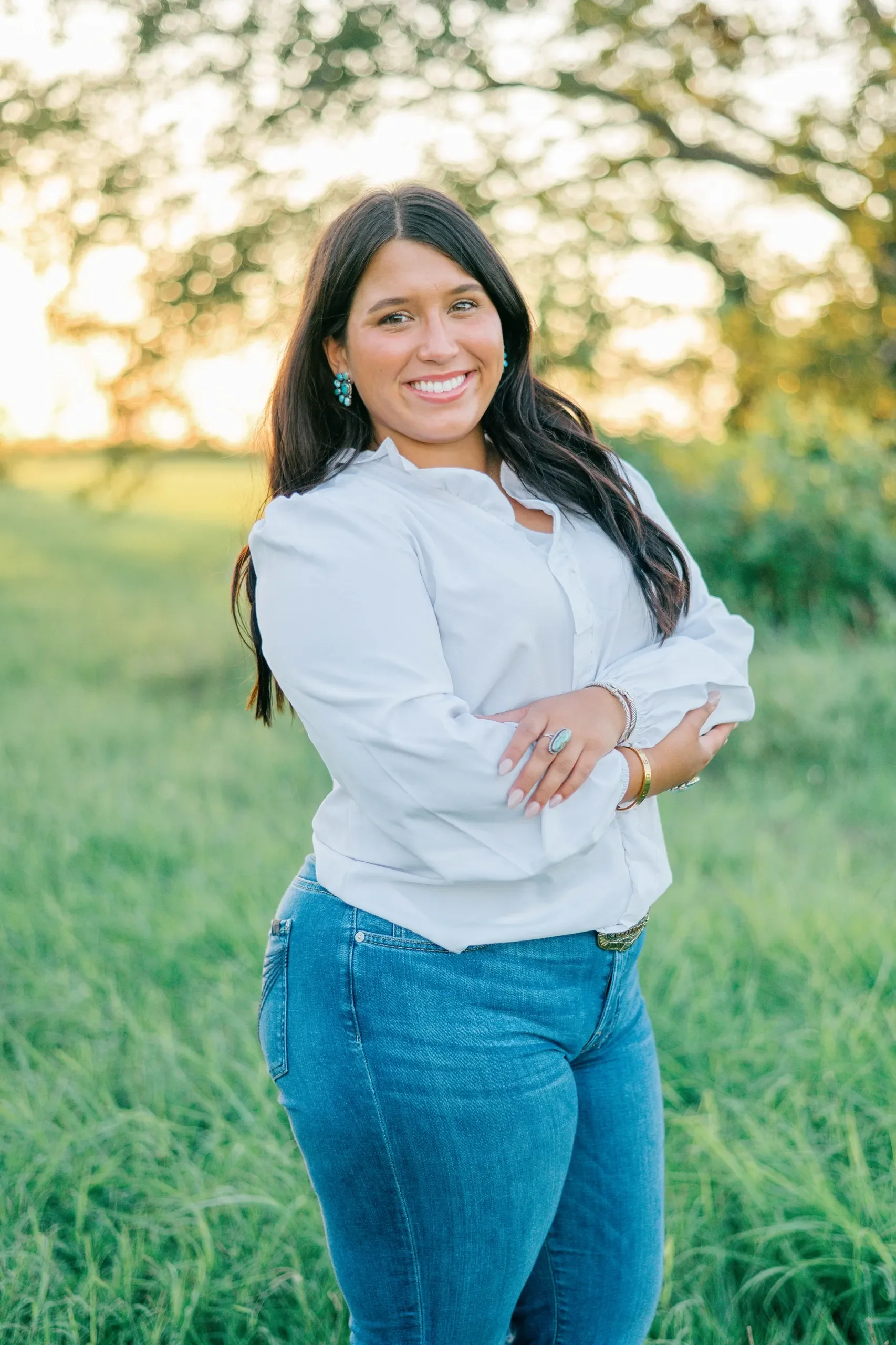 A woman with long dark hair wearing a white button-down shirt and blue jeans, smiling with arms crossed, standing outdoors in a grassy field during sunset.