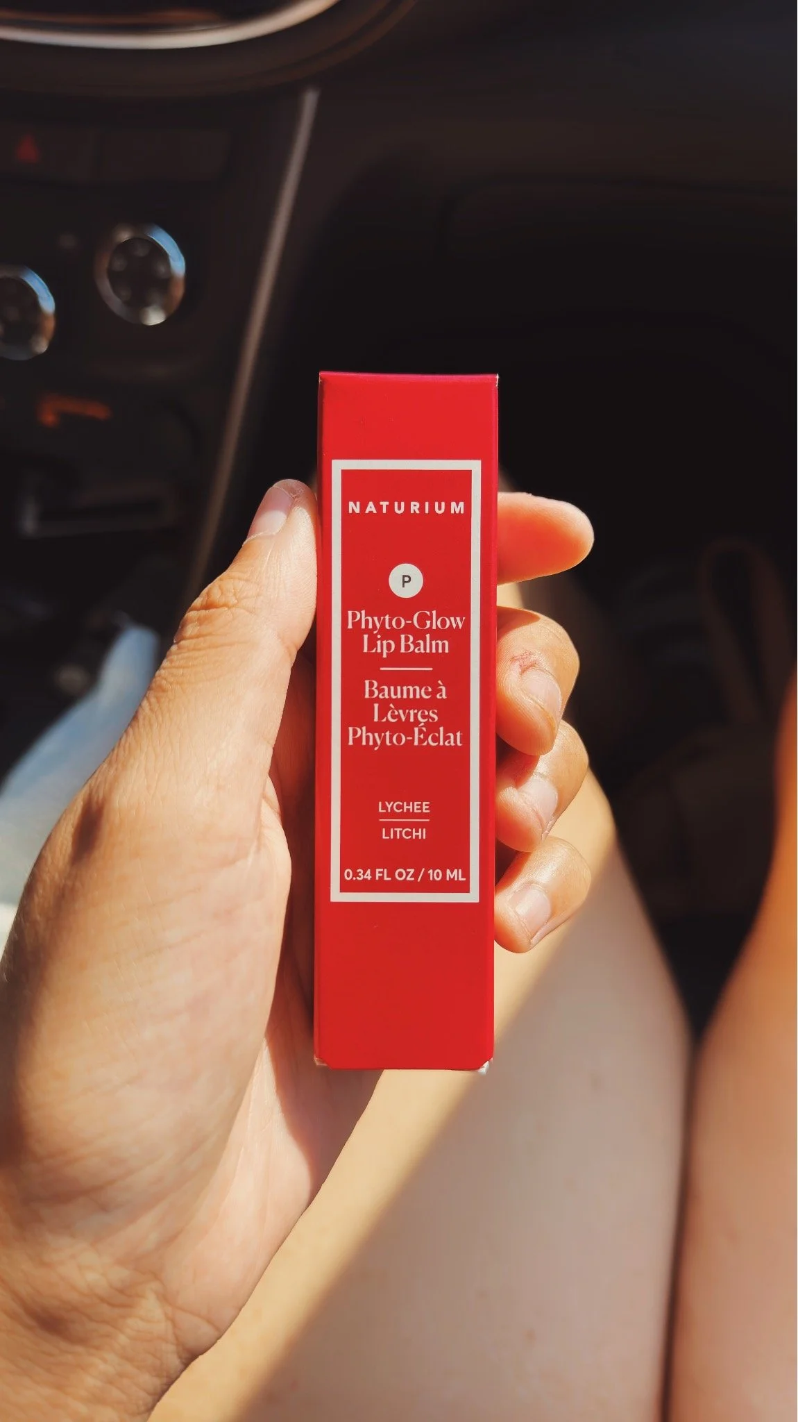 A person's hand holding a red box of Naturium Phyto-Glow Lip Balm inside a car, with the dashboard and person's legs visible in the background.