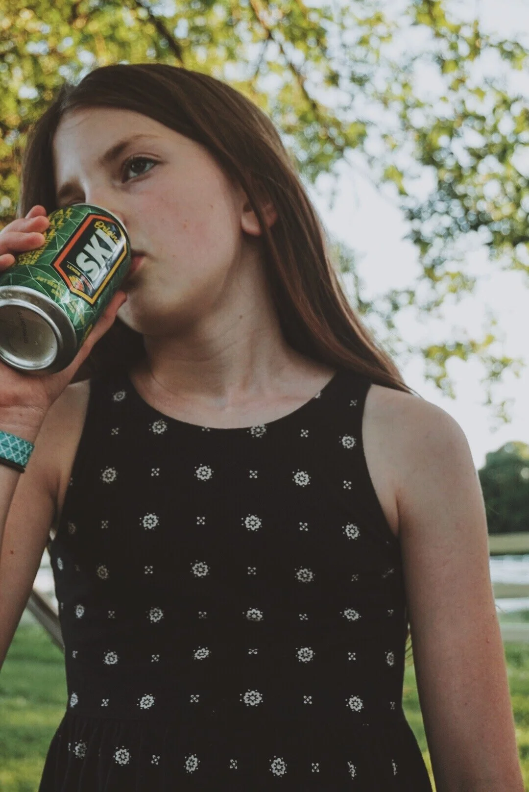 A young woman with long brown hair drinking from a can outdoors during daytime, with green trees and blue sky in the background.
