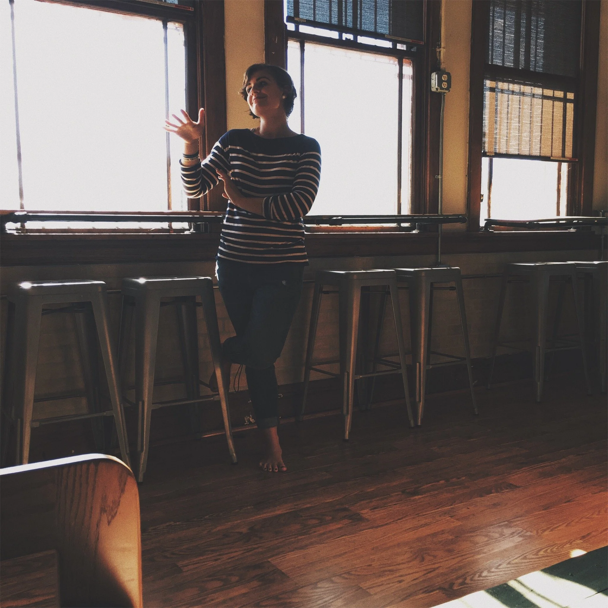 A woman standing barefoot near large windows in a room with wooden floors and bar stools, gesturing with her hands.