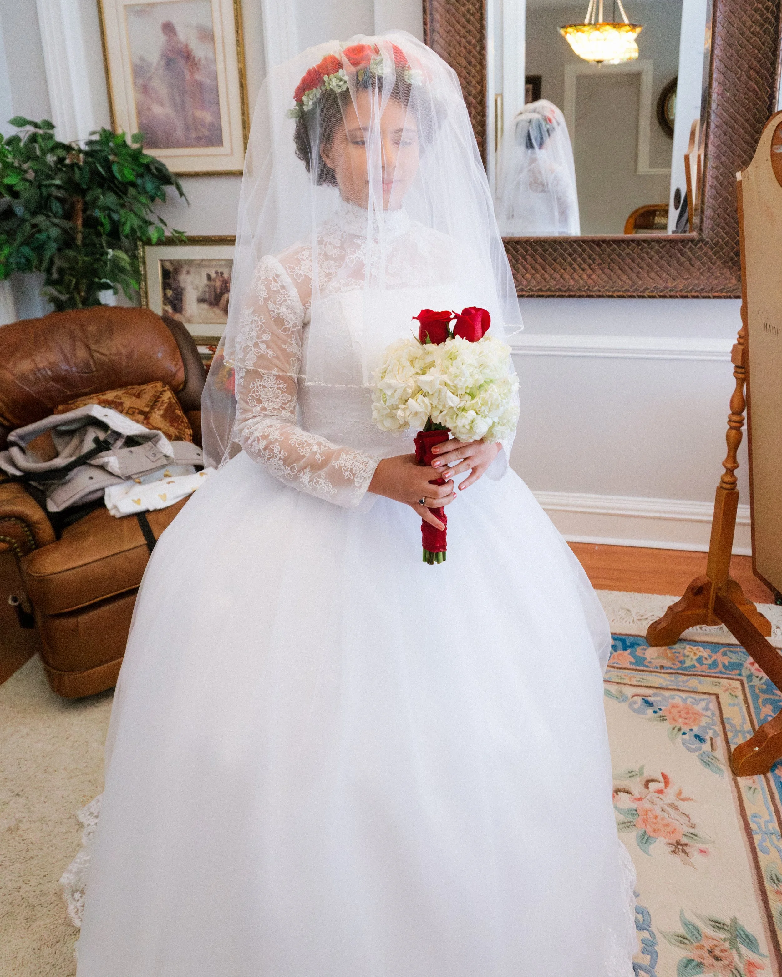 A bride in a white wedding dress holding a bouquet of white and red flowers, standing indoors in front of a mirror and a framed picture.