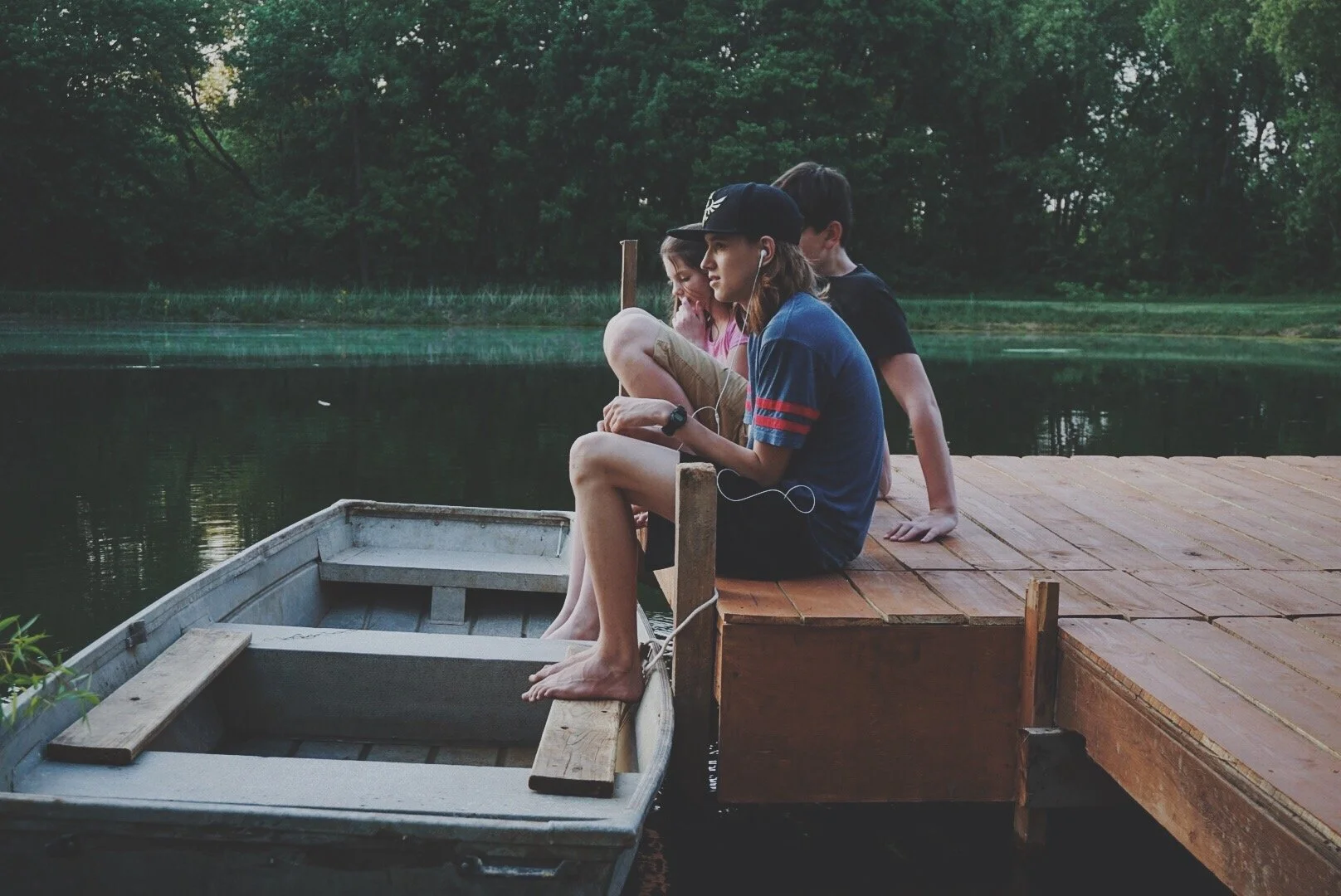 Three kids sitting on a wooden dock by a lake, with a small rowboat in front of them, surrounded by trees.