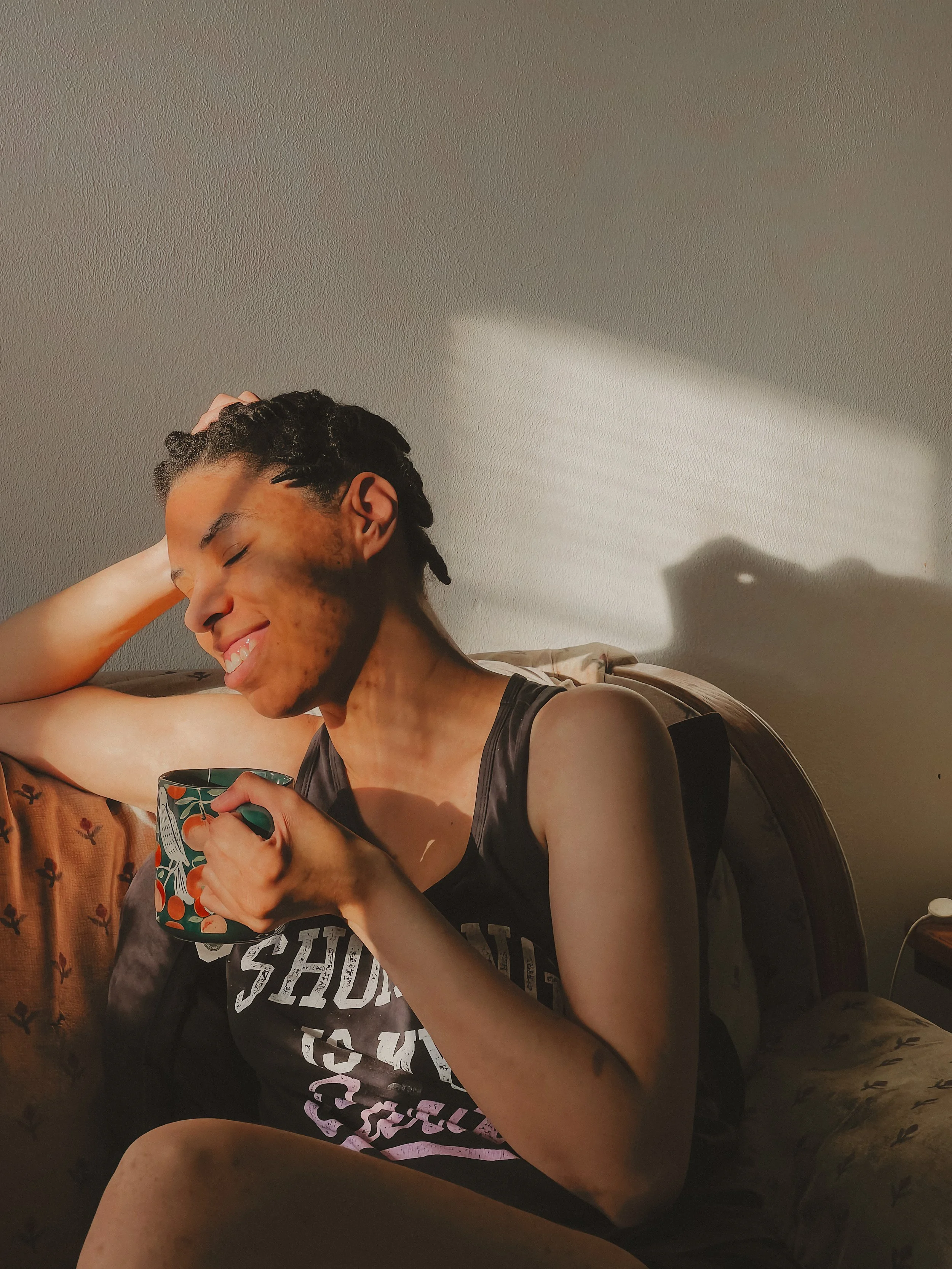 A young man with dreadlocks sitting on a sofa, smiling and holding a colorful mug. He is wearing a sleeveless black shirt and appears relaxed, with sunlight casting shadows on the wall behind him.
