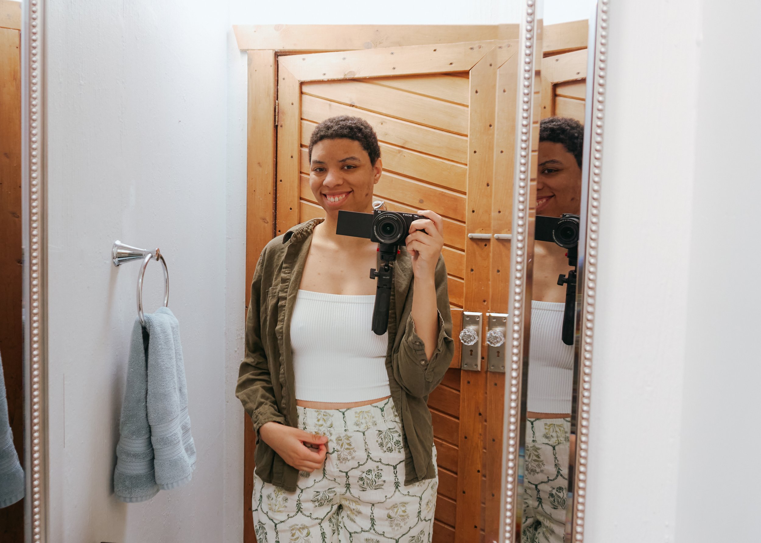 Young person smiling while taking a mirror selfie with a camera in a bathroom with wooden door and white wall.