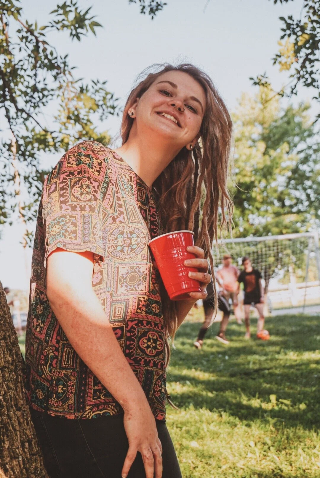 A young woman with a big smile, holding a red cup, standing next to a tree in a park, with a soccer game in the background.