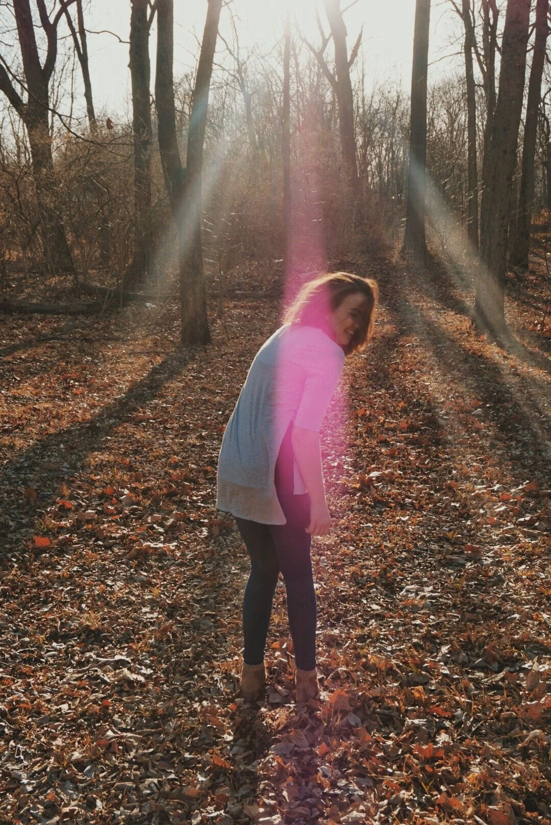 A woman laughing while standing in a forest during autumn, with sunlight streaming through the trees.