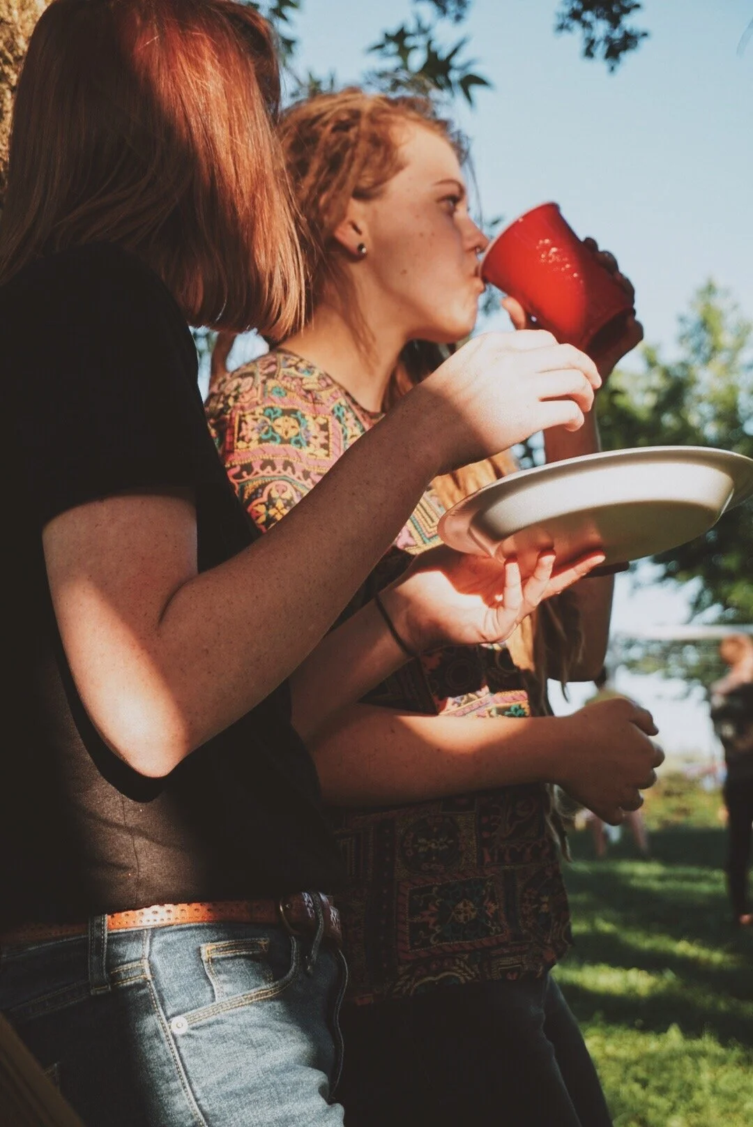 Two young women with red hair are standing outside, one drinking from a red cup while holding a white plate. They are in a green, sunny environment, possibly at a picnic or outdoor gathering.
