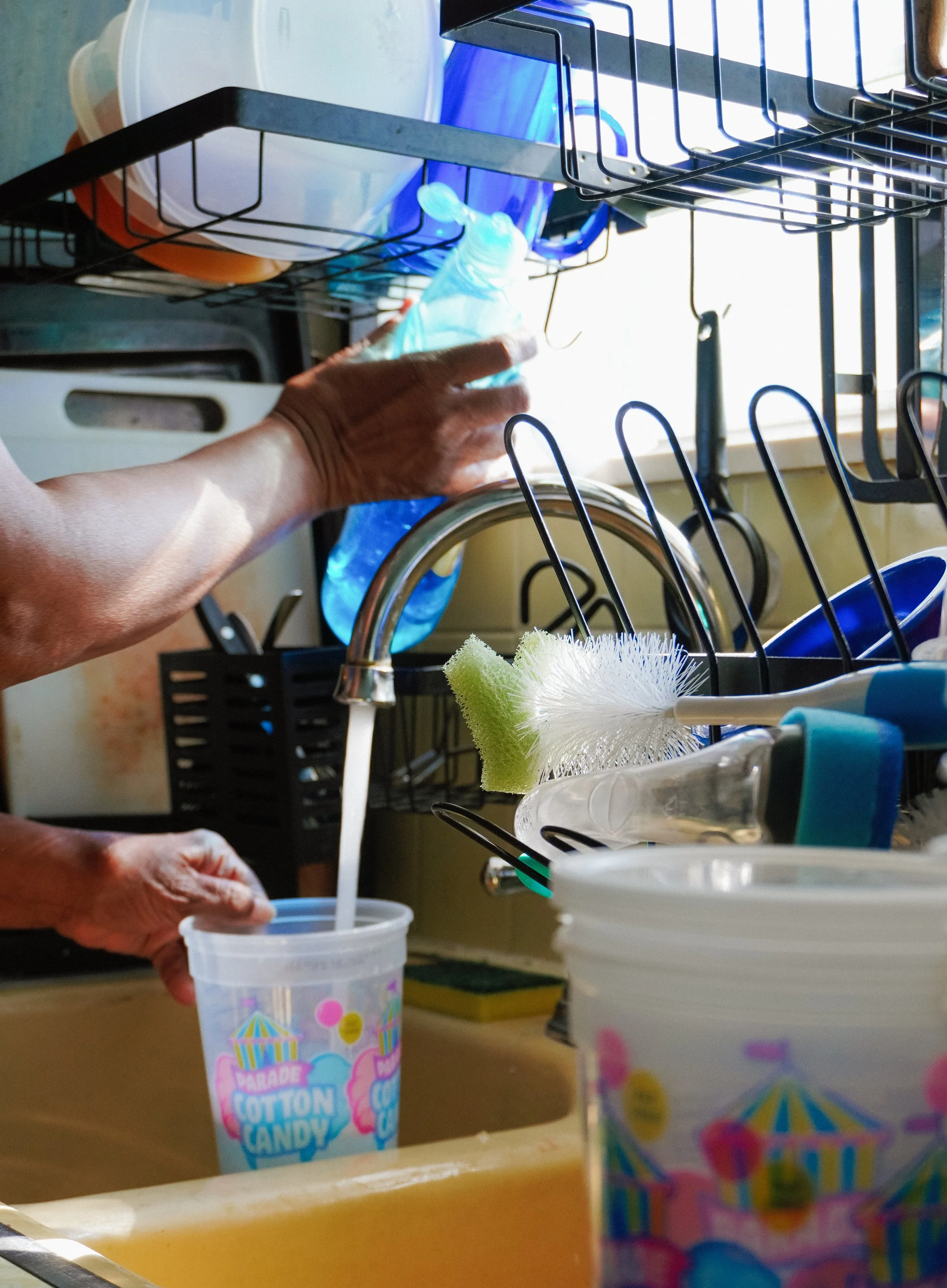 Person filling a blue cup with water at a kitchen sink, with dishwashing tools and containers nearby.