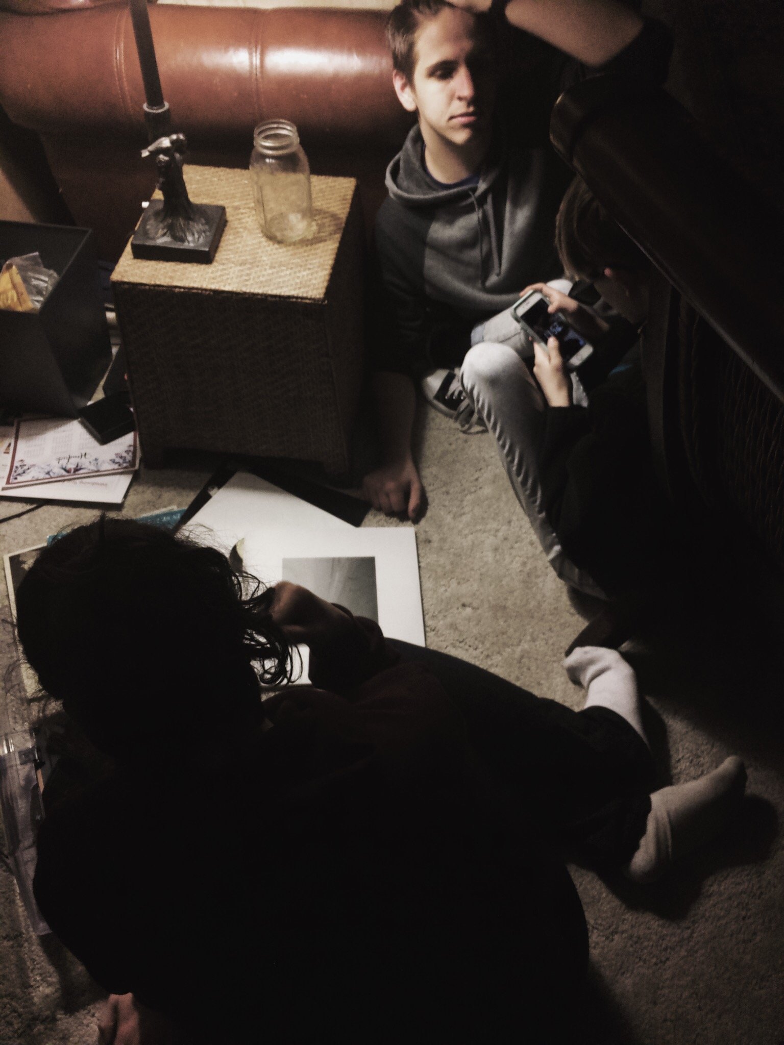 Three people sitting on a carpeted floor with one using a smartphone, one sitting next to a small table with a jar and a lamp, and the third person sitting on the floor looking at a photo print.