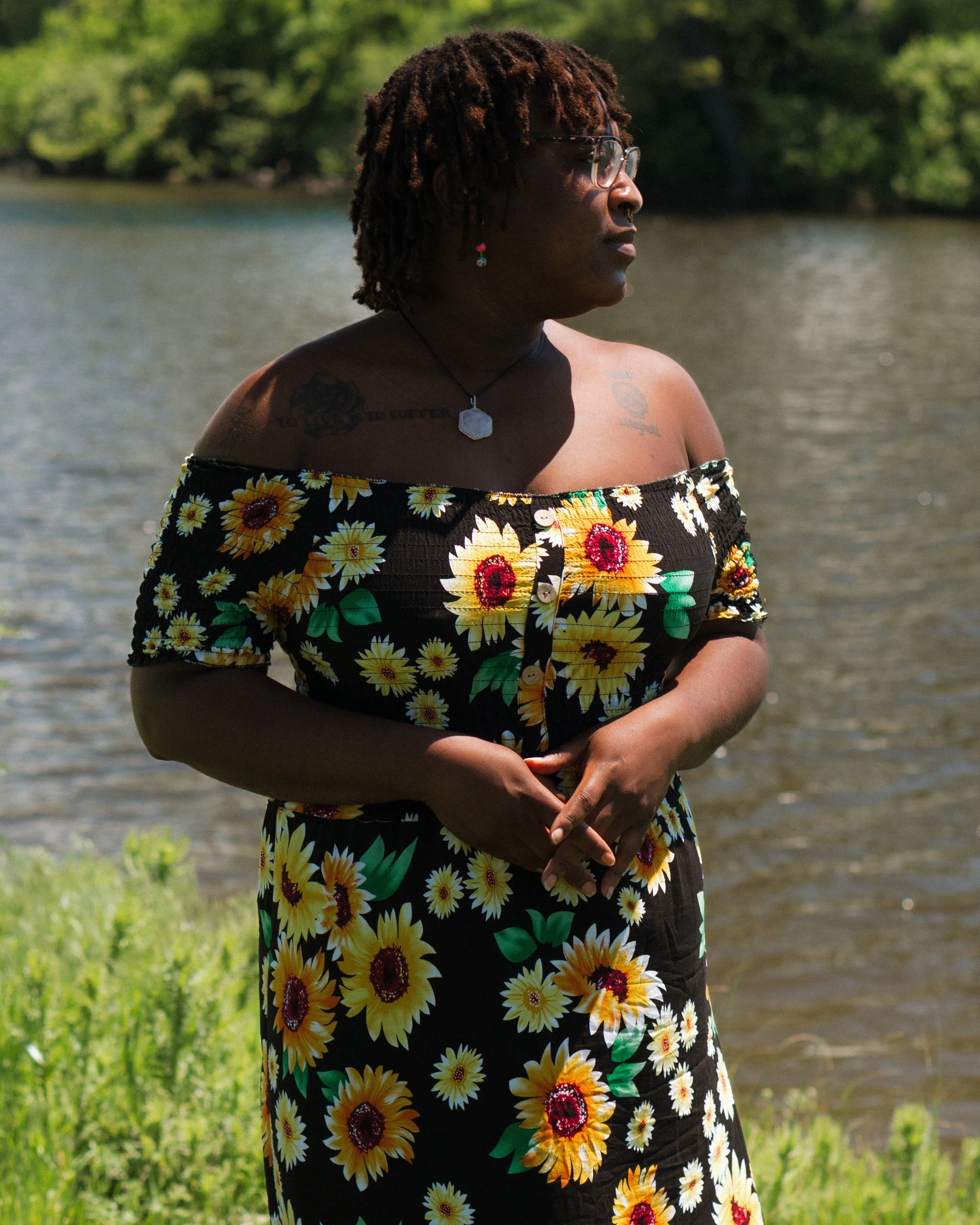 A woman standing by a body of water, wearing an off-shoulder black dress with a yellow sunflower print, sunglasses, and earrings, with green trees in the background.