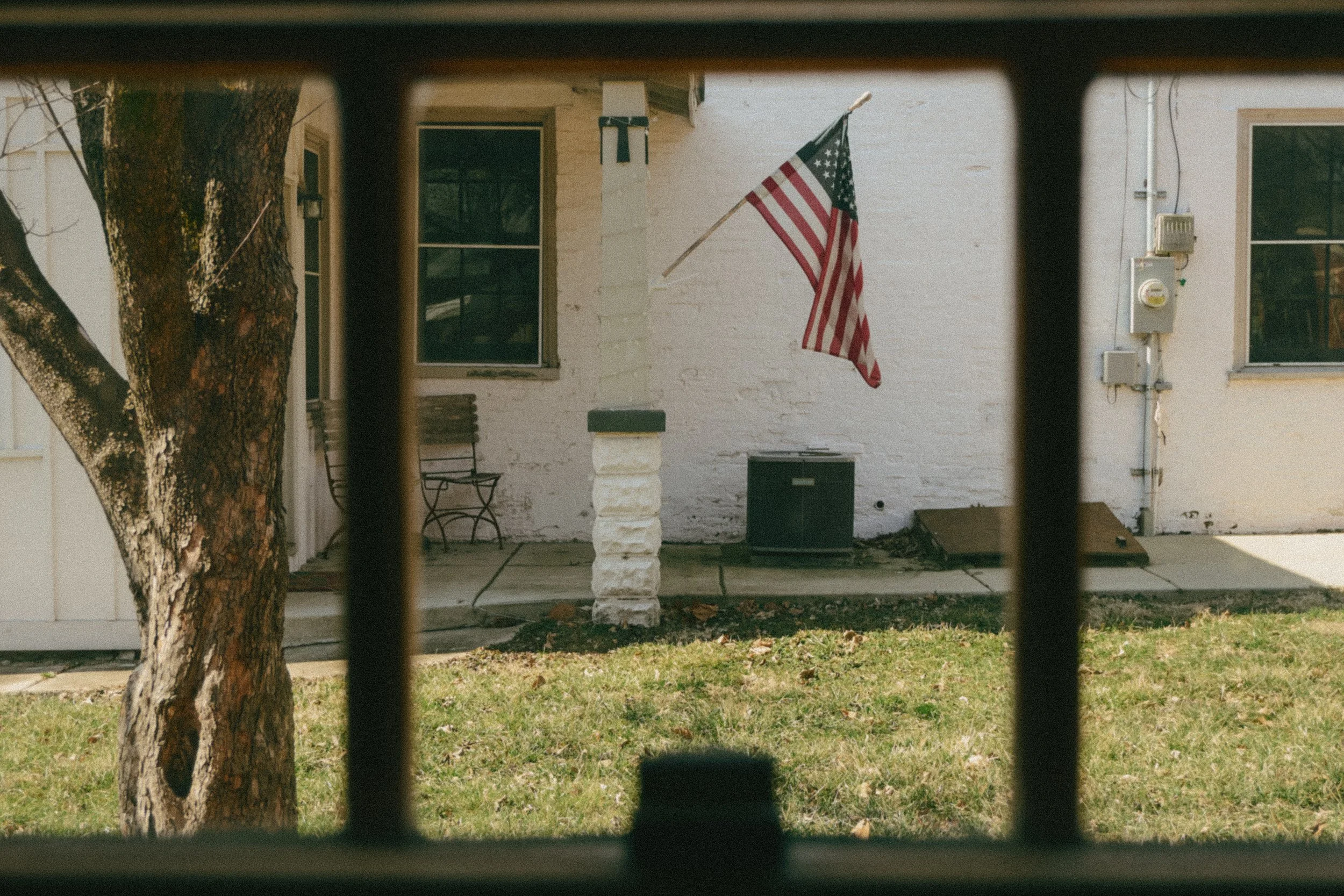 View of a house porch with American flag mounted on the wall, a wooden bench, and a black object on the ground, seen through a window with metal bars.