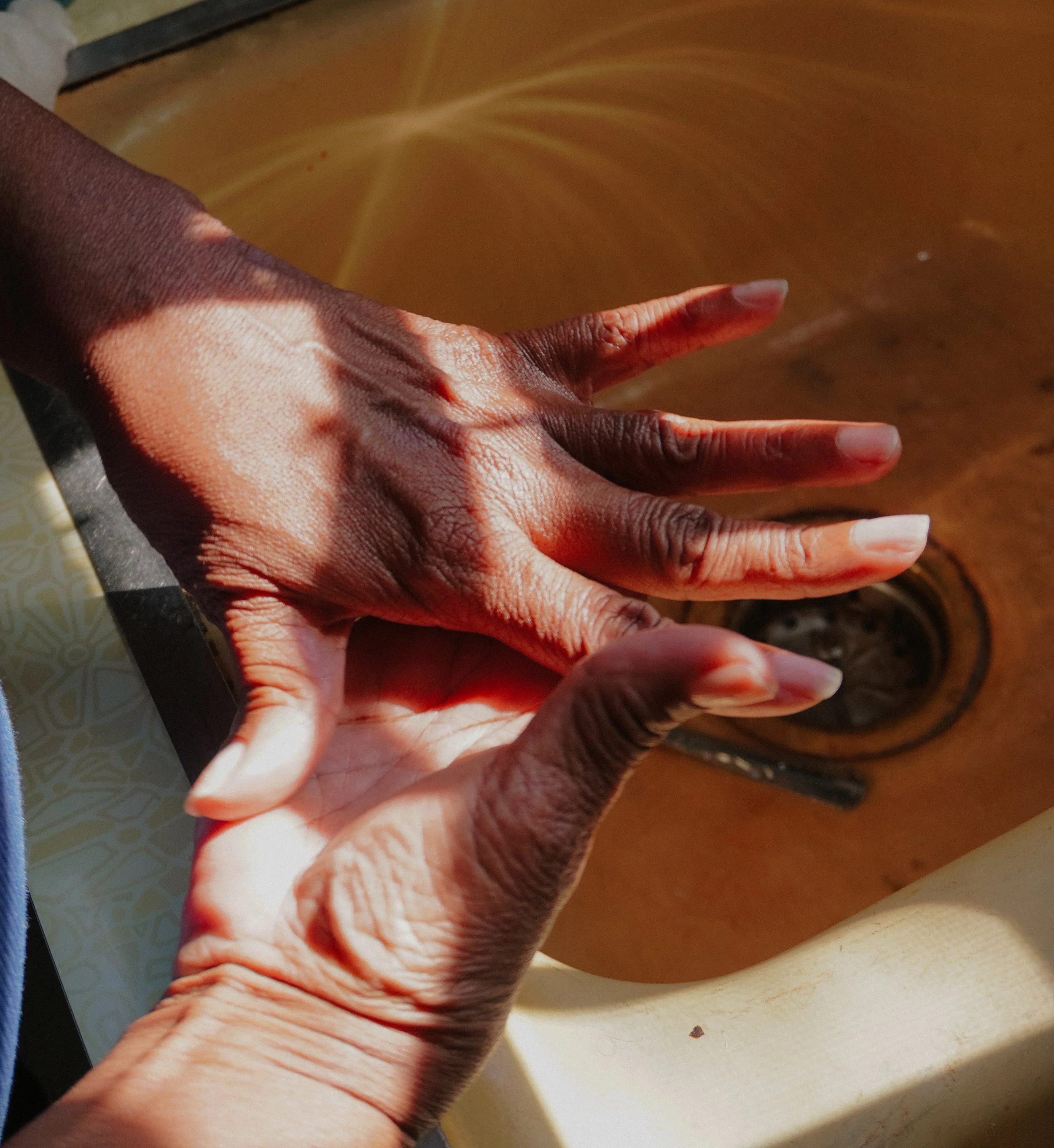 A pair of hands with dark skin are washing, with sunlight casting shadows, over a yellow sink with a water drain.