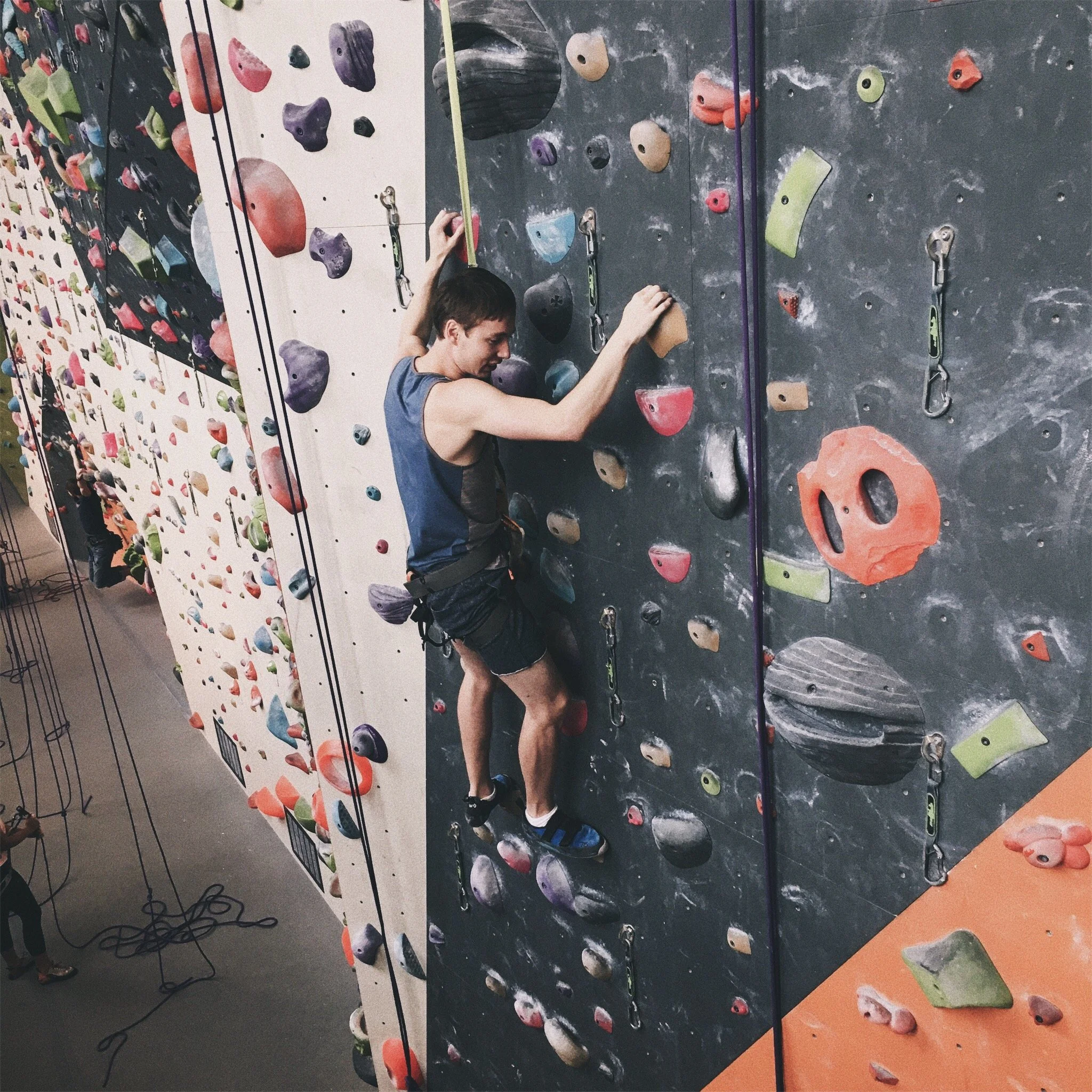 A young male climber scaling an indoor rock climbing wall with colorful holds.