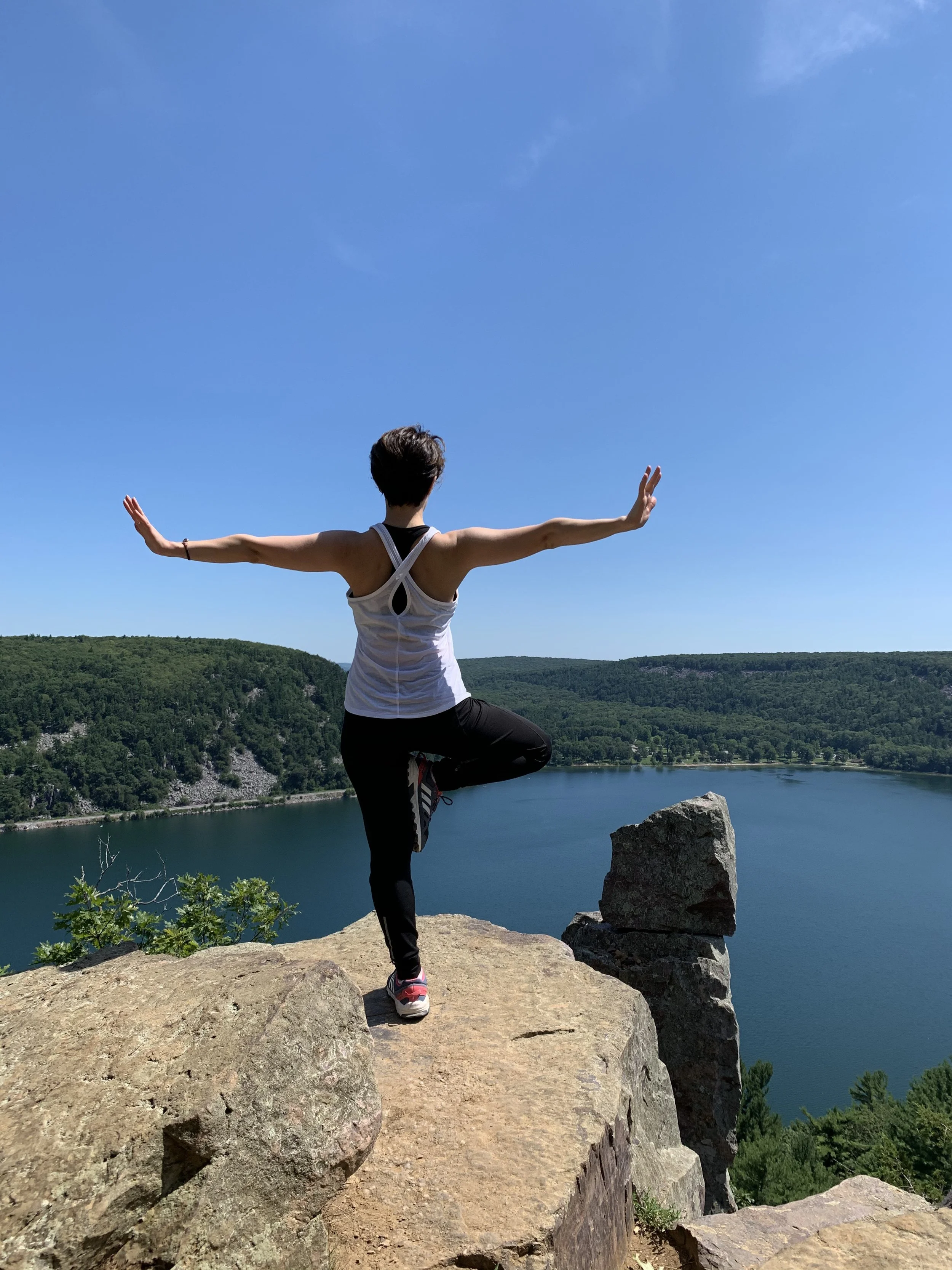 A woman practicing yoga on a rocky ledge overlooking a lake and green hills under a clear blue sky.
