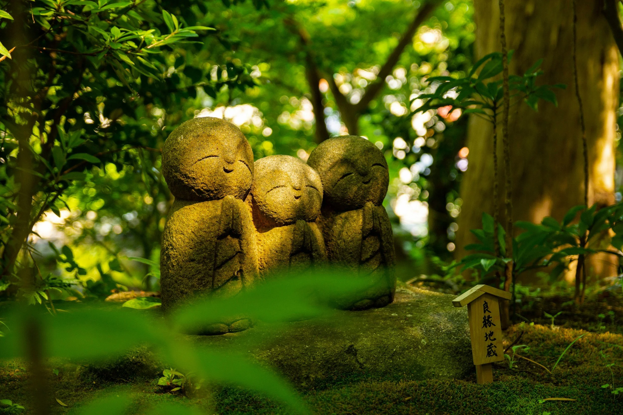 Three small stone statues of monks with closed eyes and peaceful expressions, standing close together in a green forest setting, with sunlight filtering through the leaves.