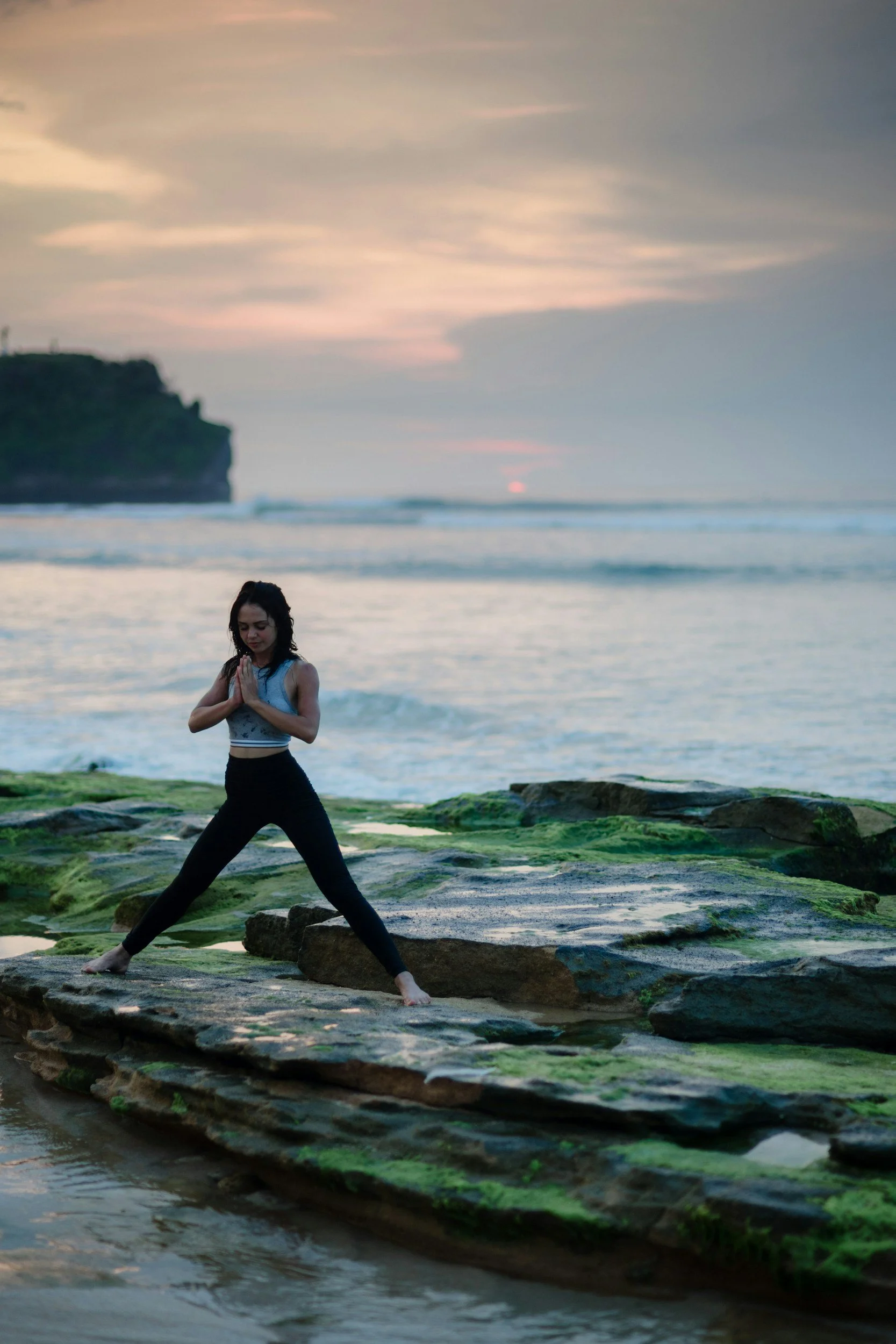 A woman practicing yoga on moss-covered rocks at the beach during sunset.