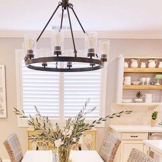 A chandelier hanging above a dining table with a vase of flowers in a bright, modern kitchen with white cabinets and open shelves.