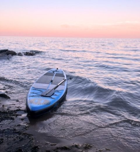 Stand-up paddleboard resting on a rocky shore at sunset, with calm ocean waters and a pink and orange sky.