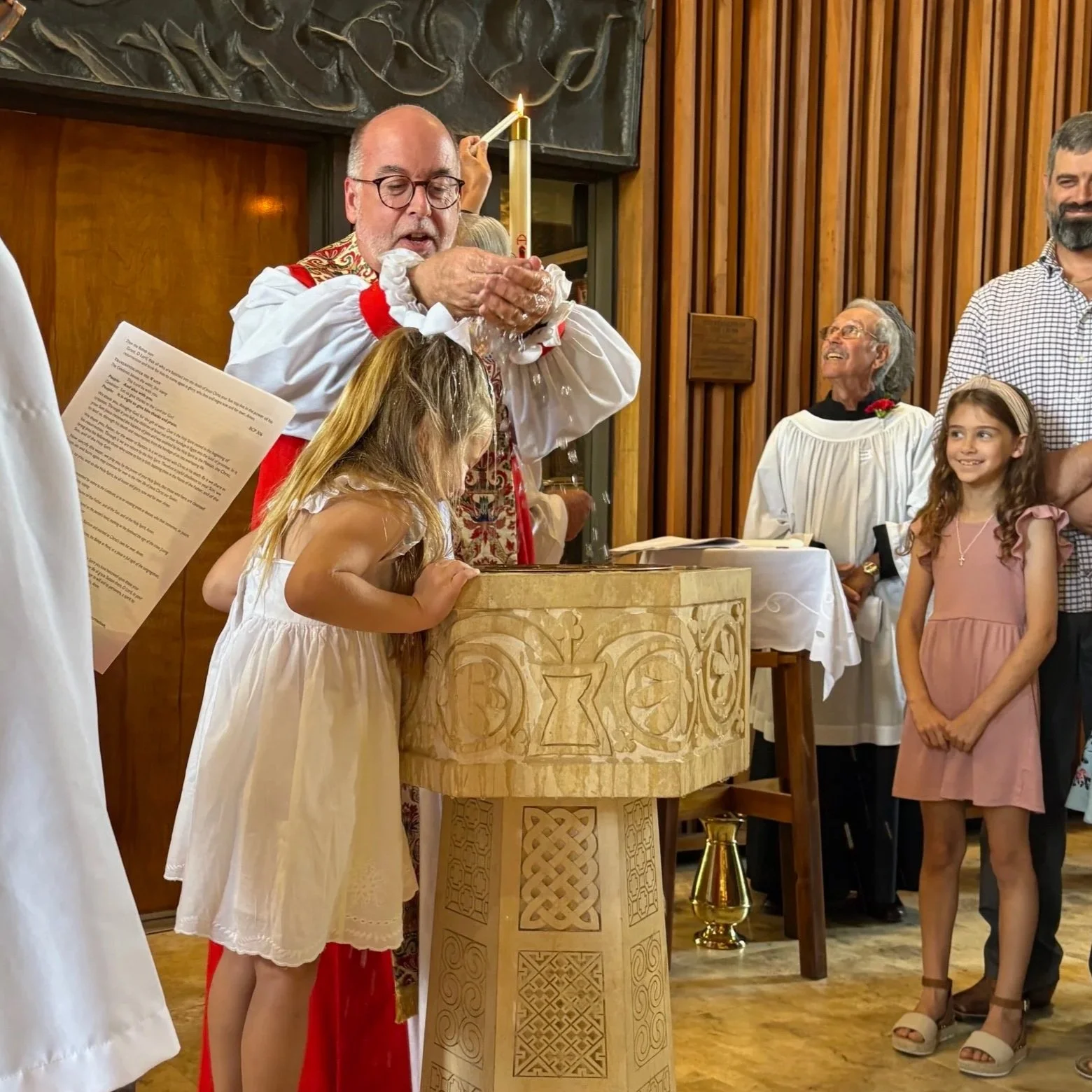 A bishop performing a child baptism at the stone baptismal font inside the sanctuary of St. Stephen's Episcopal Church, with family members gathered around watching.