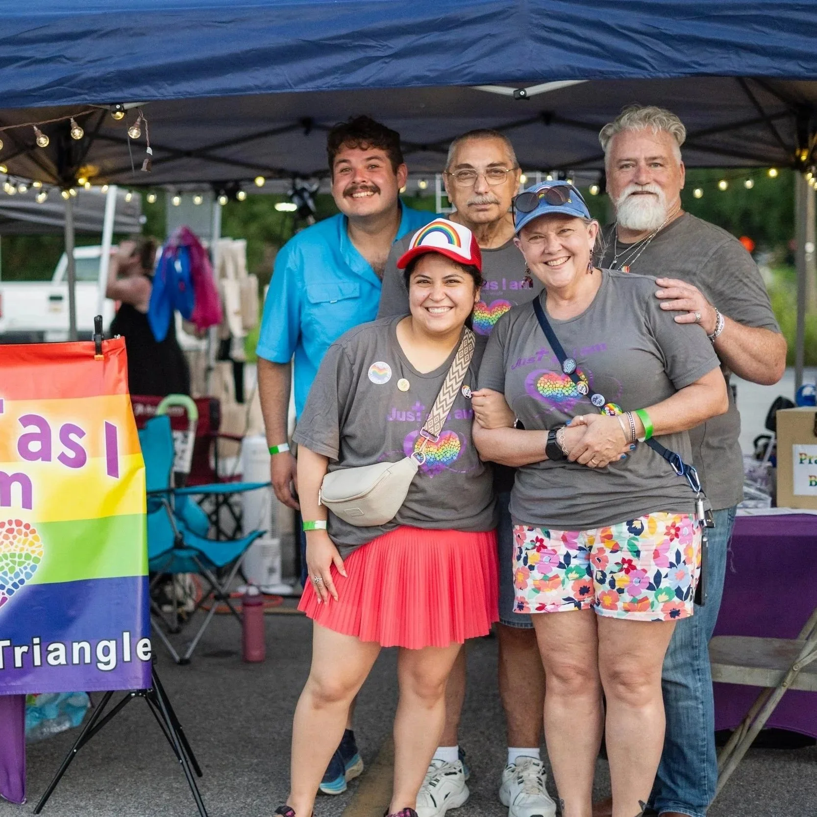 A group of smiling missional community members standing together outdoors under string lights doing outreach at the local Pride Fest event..