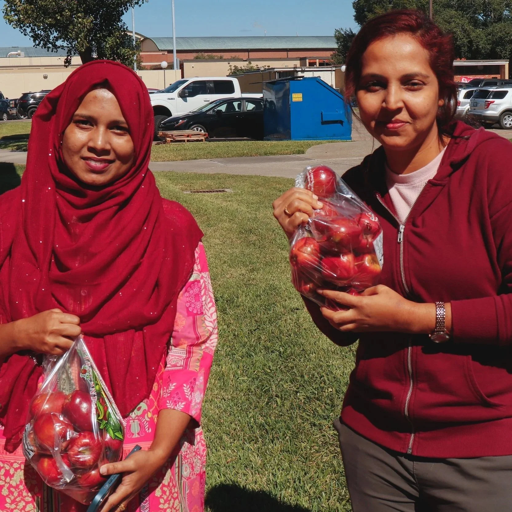 Two smiling women holding bags of fresh red apples at a community food outreach event hosted by St. Stephen's Episcopal Church, St. Mark's Episcopal Church, and The Wesley Foundation at Lamar University..