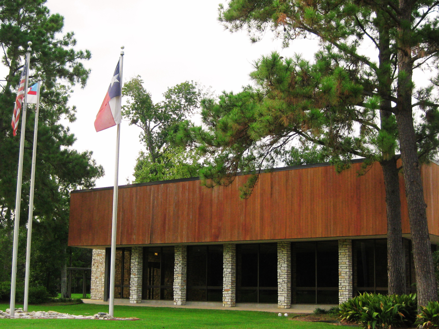 A mid-century modern building with stone columns and a wooden fascia, surrounded by green trees and grass, with three flagpoles in front flying the American flag, Texas flag, and another flag.