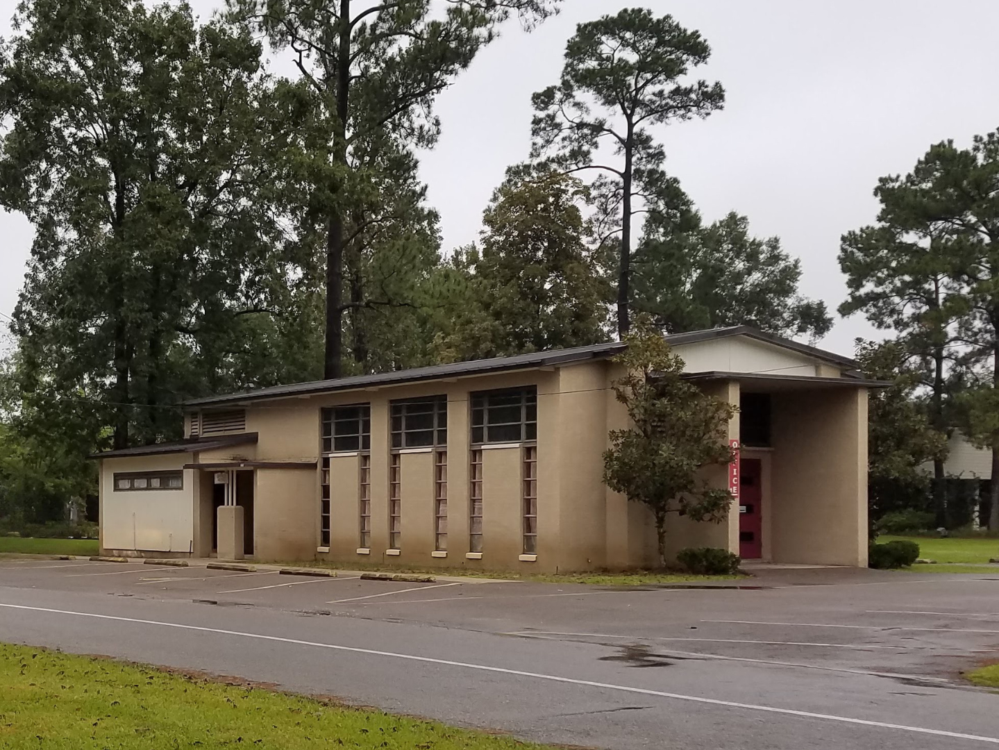 A beige, mid-century modern building with tall narrow windows, and a tree in front, situated in a parking lot surrounded by green grass and trees.