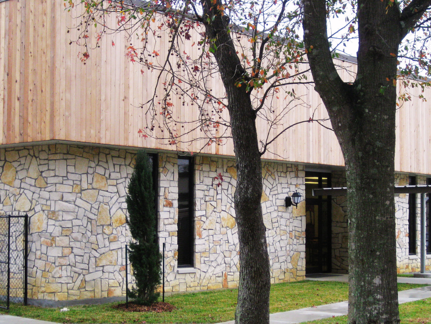 A modern building with a stone facade and wooden upper section, surrounded by grass and trees with sparse leaves.