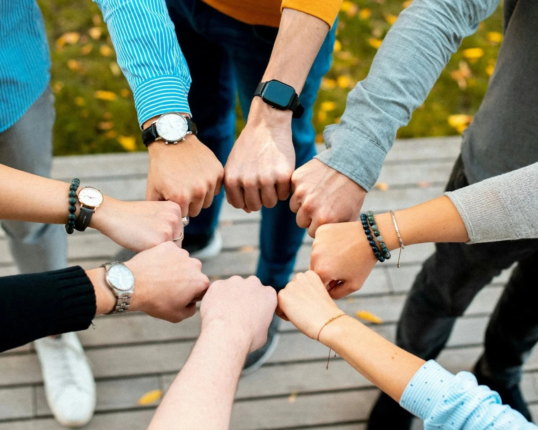 Group of diverse people wearing watches and bracelets, joining hands together in a circle on a wooden boardwalk. The background shows trees with autumn leaves.