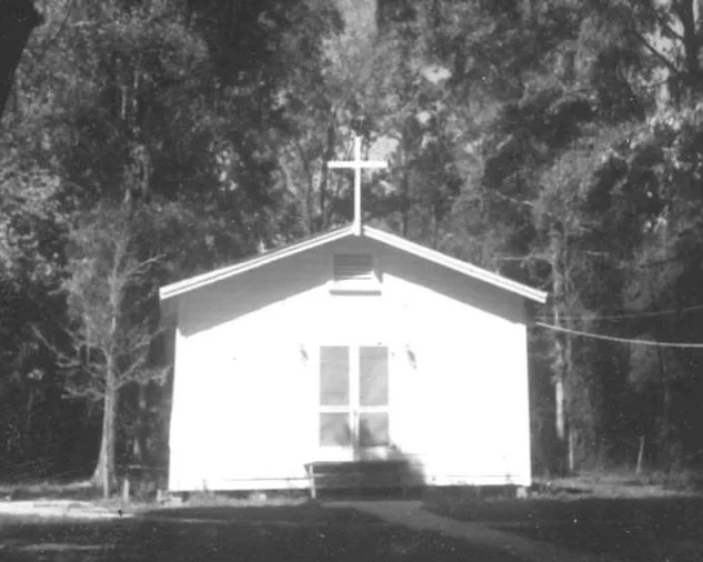 A white wood frame building with double screen doors on the front and a cross at the apex of the roof. It is a pier and beam structure on a partially wooded lot.