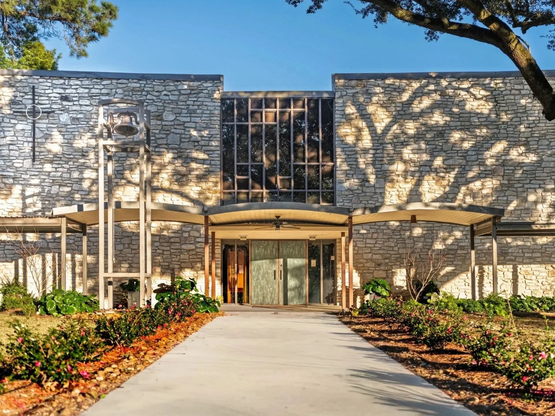A modern building exterior made of light-colored stone with large glass windows at the top, surrounded by a landscaped garden with bushes, flowers, and trees, and a concrete pathway leading to the entrance.