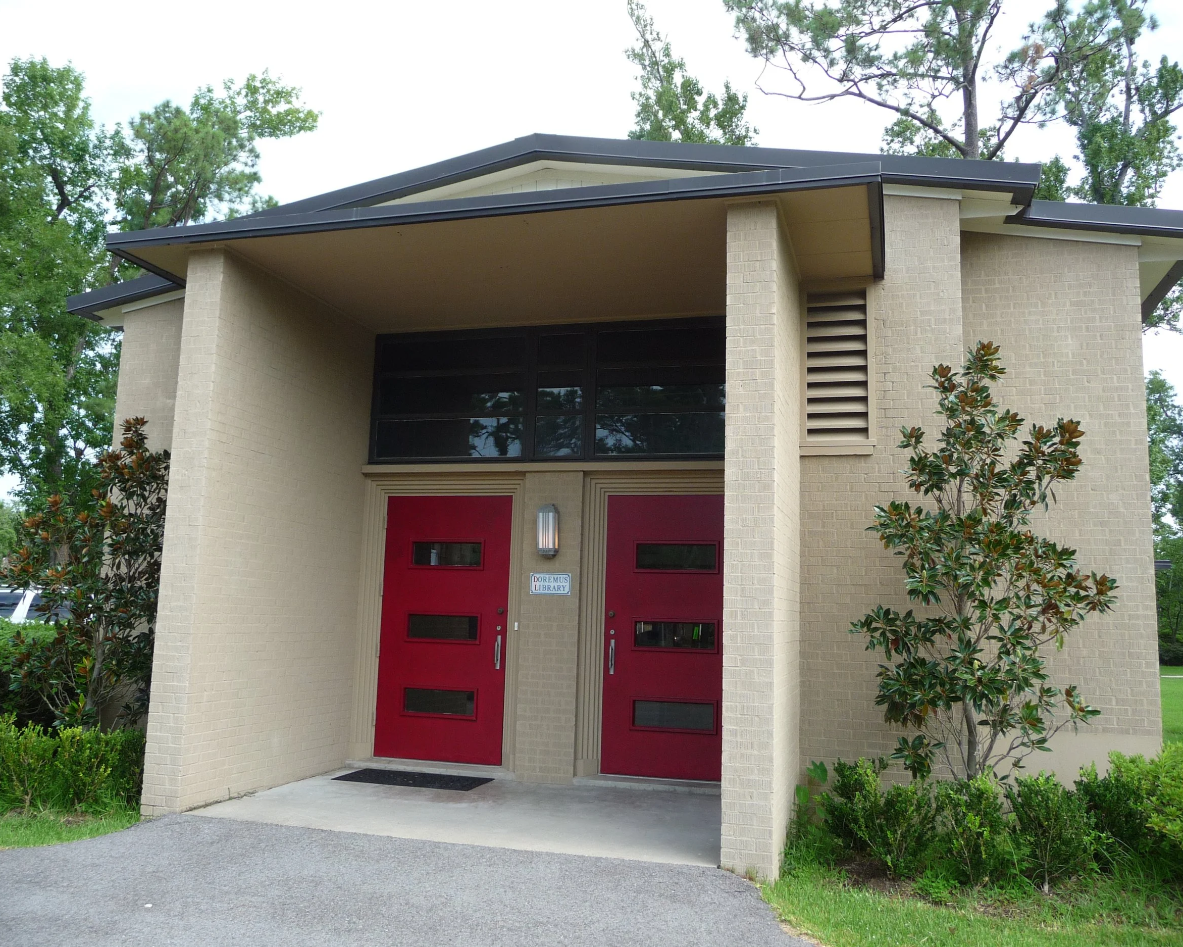 A tan building with double red doors on the front and greenery on either side of the porch.